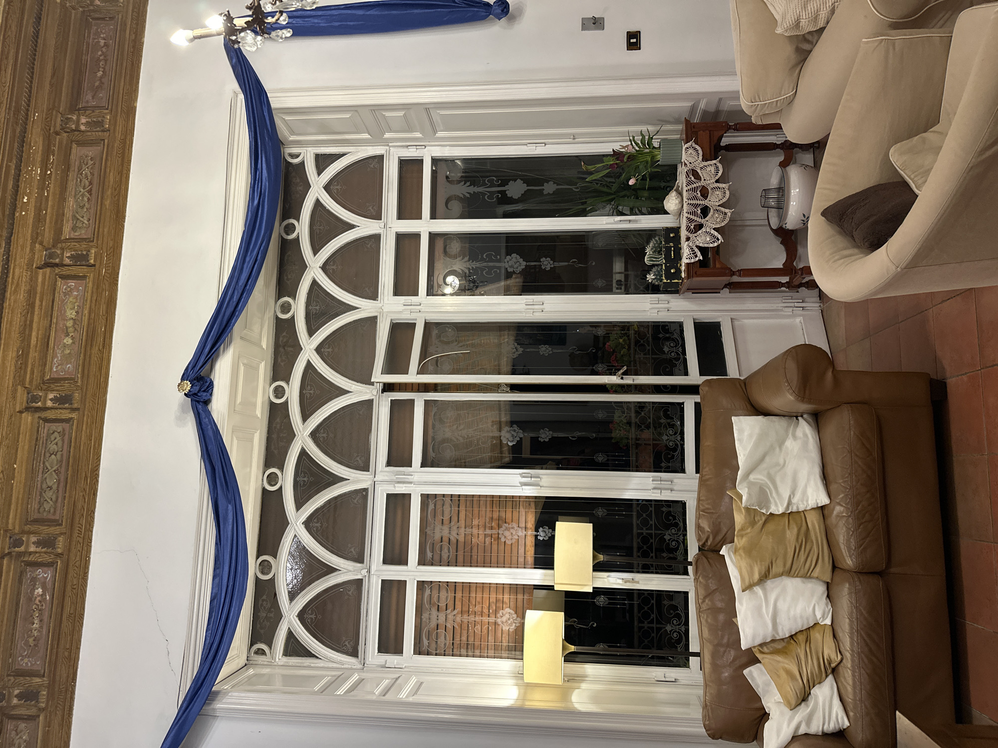 The photograph shows a section of the interior of a space with a large glass door decorated with a white frame. Draped on the left side of the door is a blue sash tied at the top. To the right is a beige loveseat and a brown sofa with various pillows. In front of the glass door are two lamps that appear to be on. Outside the glass door, in the background, are what appear to be brick-like structures, possibly buildings or a wall. Based on the presence of Italian language content (possibly implied by the mention of Catania, Italy), the scene could be in a domestic interior of a house or apartment in Catania, Italy.