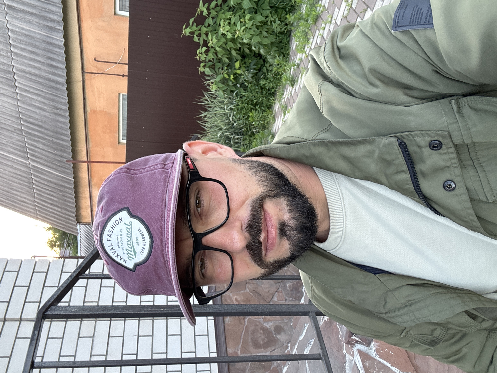 The image shows a man with a beard and glasses wearing a maroon baseball cap and a green jacket. He is standing outdoors with a brick wall to his left and a wooden fence and greenery behind him. The location appears to be a residential area, possibly a backyard, in Novaya Usman', Russia. The man is looking towards the camera, and the overall lighting suggests it is daytime. The background includes a tiled roof and a light orange wall, consistent with rural or suburban Russian architecture.