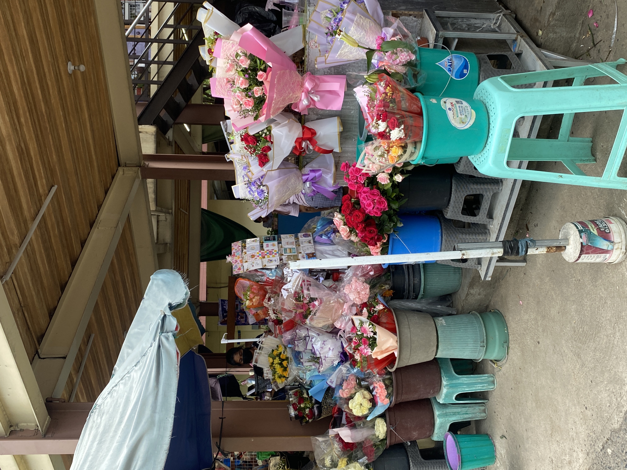 This is a full-shot, eye-level photograph of a flower stall located in the Agdao Public Market in Davao City, Philippines. The stall is filled with various bouquets of flowers, predominantly roses in shades of red, pink, and yellow, as well as white daisies and purple flowers. These bouquets are displayed in buckets and on tiered shelves. Some bouquets are wrapped in transparent plastic and decorative paper, tied with ribbons. In the background, a person wearing a black face mask is visible behind the stall, seemingly attending to the flowers. The stall is covered by a light blue awning, and the surrounding environment suggests an outdoor market setting with concrete flooring and a wooden ceiling structure. The overall scene depicts a vibrant flower market stall.