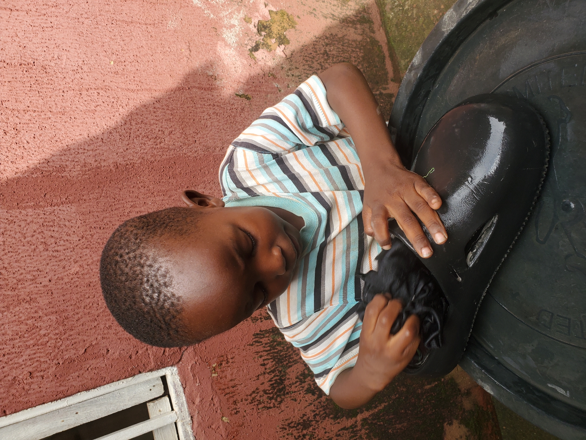 An African boy, appearing to be around 4-6 years old, is shown from a high angle, focused on cleaning a black sandal. He is wearing a striped t-shirt with horizontal bands of blue, white, orange, and black. His hands are actively engaged in wiping the inside of the sandal with a black cloth. The sandal rests on the rim of a large, dark-colored plastic basin. The background consists of a textured, reddish-brown wall. To the left of the boy, a portion of a wooden window frame with white slats is visible, set against the wall. The scene appears to be outdoors, likely in a residential area within Jos, Nigeria, given the architectural style of the wall and window. The lighting suggests it is daytime, with sunlight casting shadows on the wall.