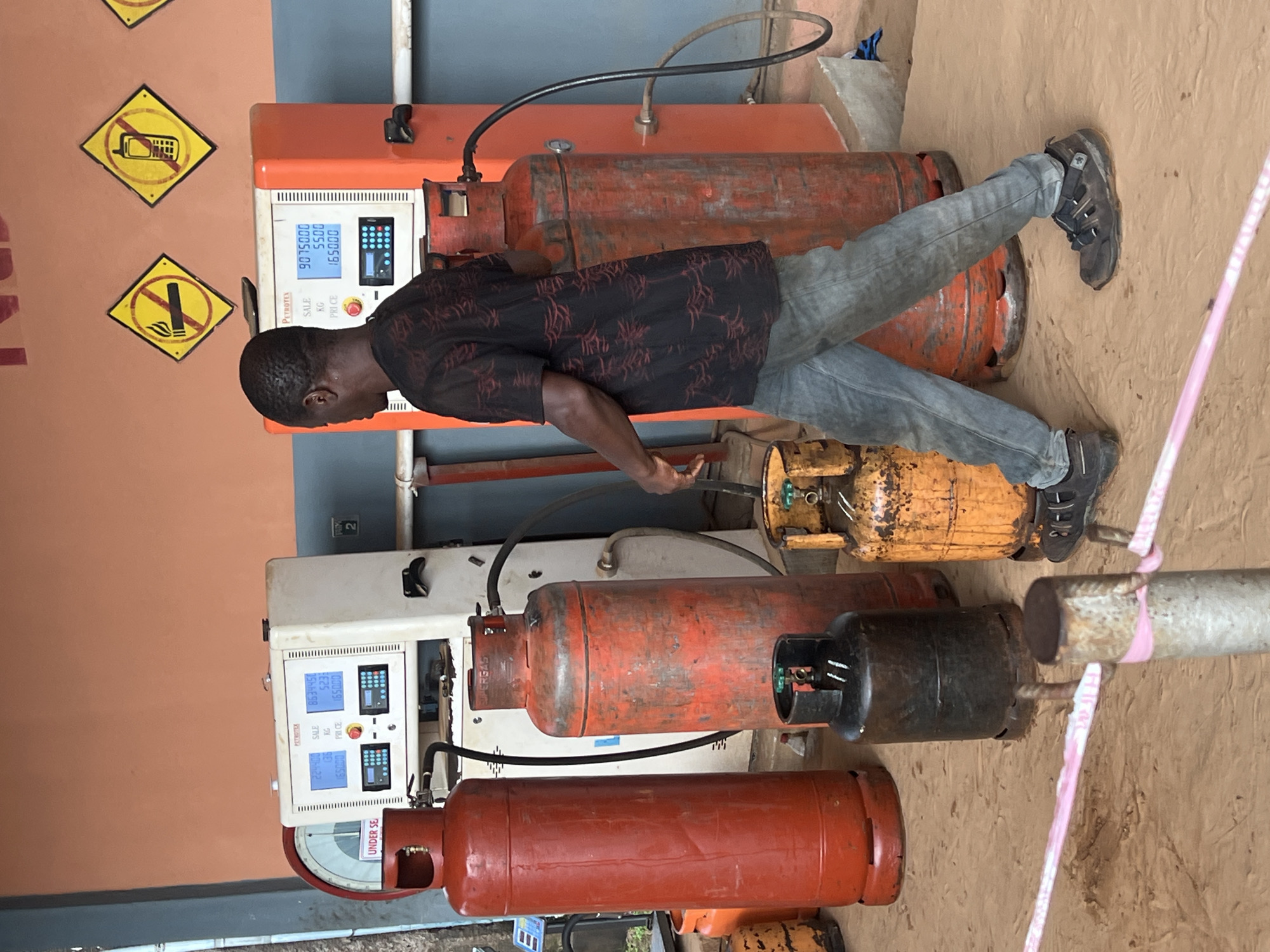 In Nkpor, Nigeria, a young man is shown working at a gas cylinder refilling station. He is wearing a black shirt with a red patterned design and blue jeans. The station is equipped with multiple gas cylinders of various colors, including orange and yellow, and two white refilling machines with digital displays. The displays show numerical values such as "9075000," "5500," "165000," "8634450," "136," and "65000," likely indicating sales, weight, and price. Two yellow warning signs are visible on the orange wall behind the man; one depicts a crossed-out mobile phone, and the other shows a crossed-out cigarette. The ground appears to be made of dirt. The overall scene suggests a typical day at a local business in Nigeria, with the man engaged in the task of refilling gas cylinders. The image is captured in an outdoor setting, possibly during daylight hours. The man's focus on his task and the orderly arrangement of equipment imply a functional and operational environment.