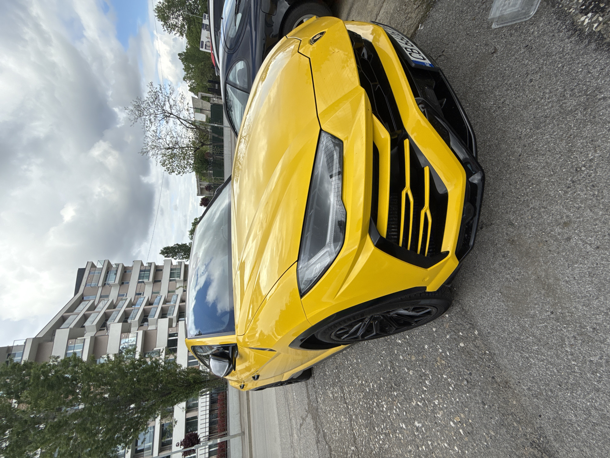 The image shows a bright yellow Lamborghini Urus parked on a street with a sidewalk in front of a modern apartment building. The car is positioned close to the curb, and another, darker-colored vehicle is partially visible behind it. The building has a unique architectural style with alternating protruding and recessed sections. The sky is overcast with various cloud formations. Trees and other greenery are present, adding a touch of nature to the urban scene. The pavement appears to be asphalt. The location is Sofia, Bulgaria.