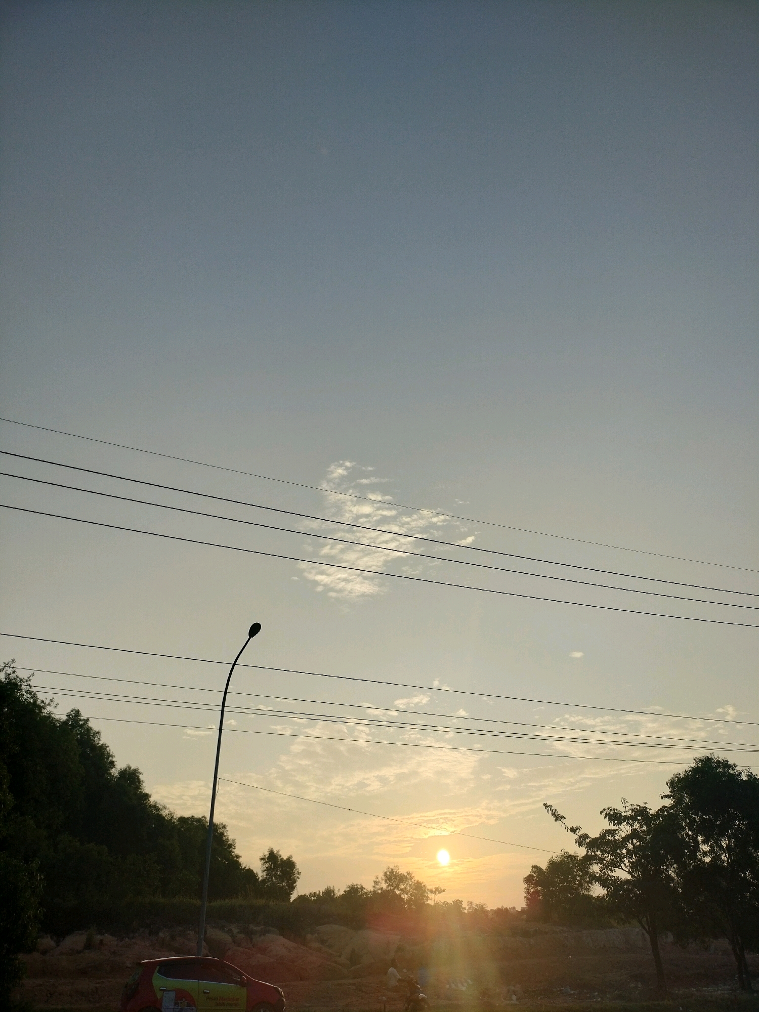 The image captures a sunset scene in Batam, Indonesia, likely along a roadside with some construction or undeveloped land. The sky is a pale blue, transitioning to a warmer yellow-orange hue near the horizon where the sun is setting. A few wispy clouds are scattered across the sky. Four parallel power lines traverse the mid-section of the image horizontally. On the left side, a dense line of dark green trees forms a backdrop, partially obscuring what appears to be a sloped embankment. A solitary street lamp with a curved pole stands prominently in the foreground on the left.
In the lower portion of the image, a red and green car is parked. To the right of the car, two figures are visible, one on a motorcycle and another standing beside it, suggesting they are engaged in a brief stop. Further to the right, more trees, silhouetted against the setting sun, are visible. The ground in the foreground appears to be a mixture of dirt and scattered debris. The overall impression is a peaceful, everyday moment captured during the twilight hours.