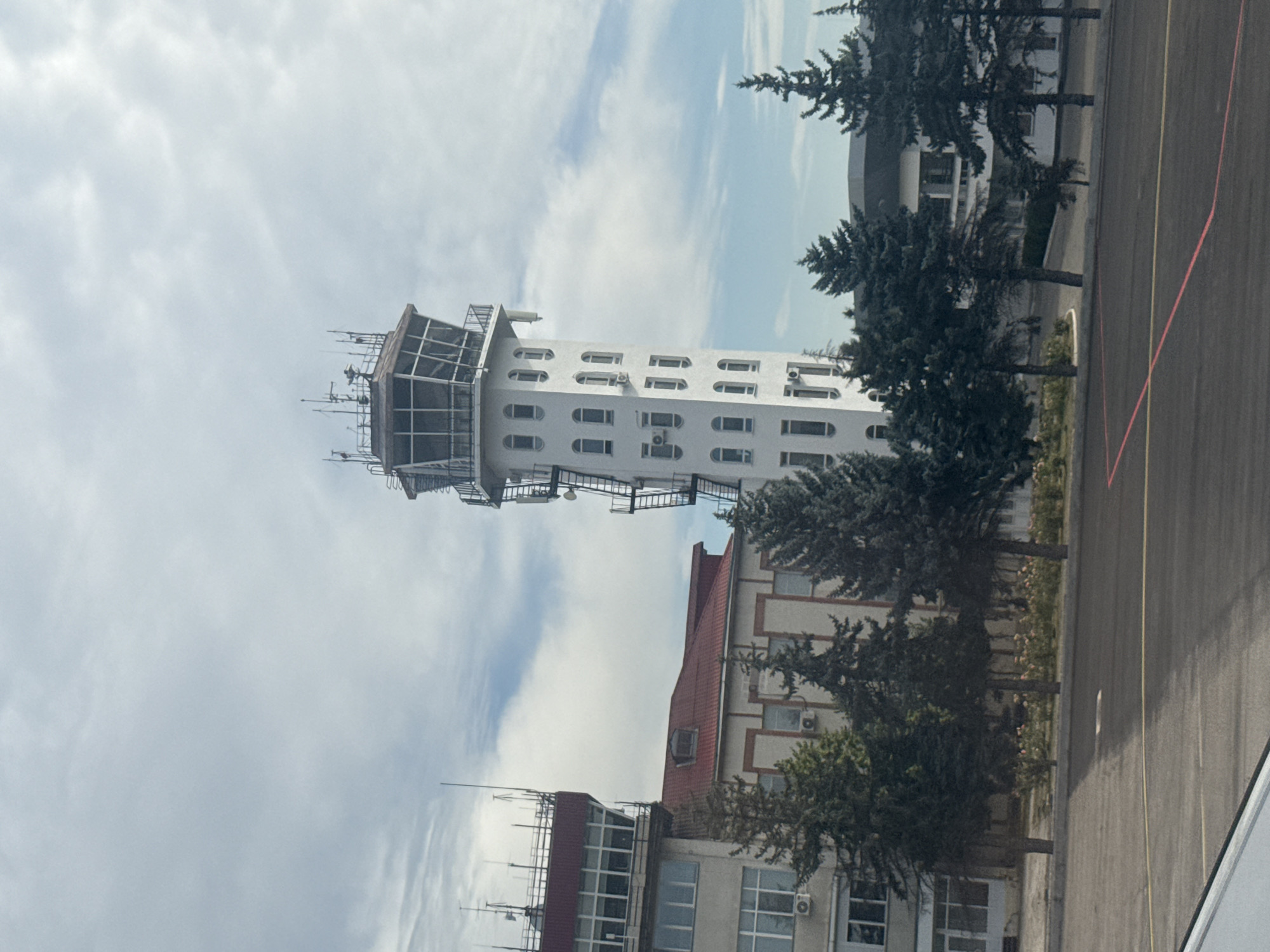 The image shows the Chișinău International Airport air traffic control tower and surrounding buildings, with a cloudy sky overhead. The main control tower is a tall, white, cylindrical structure with a glass-enclosed observation deck at the top. There are multiple windows on the tower, and an external staircase runs up its side. To the left of the main tower is a lower, rectangular building with a red roof and a control room area on top. Several evergreen trees are planted in front of the buildings. The foreground shows a dark asphalt surface, possibly a tarmac or parking area, with yellow and red markings. The overall scene depicts an operational airport environment.