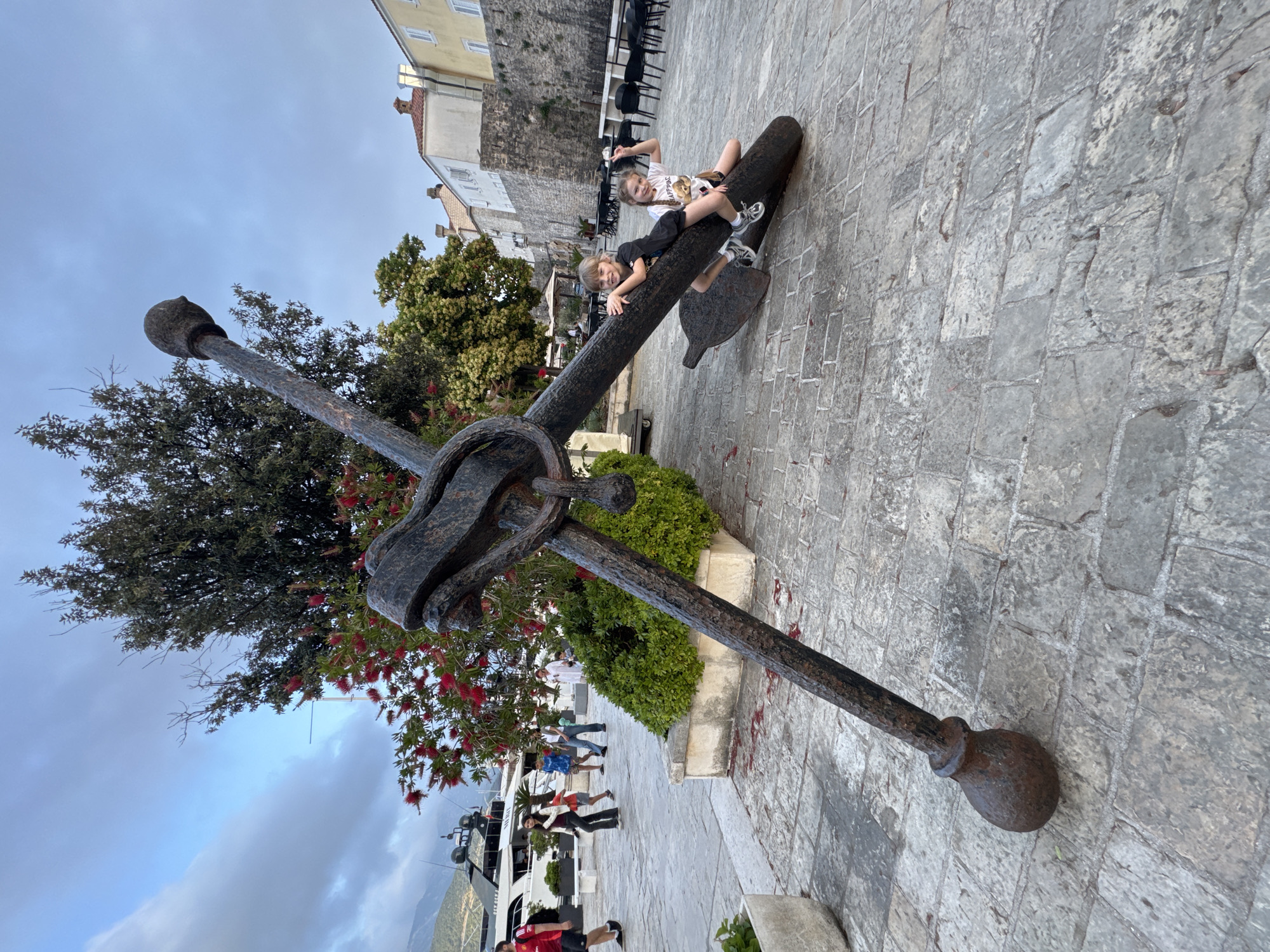 Two children are sitting atop a large, ornate metal anchor displayed on a stone-paved area. The anchor is situated near trees and flowering shrubs. Behind the anchor, a building is visible in the distance. Other people can be seen walking in the background. The location is in the city of Budva, Montenegro, as inferred from the surrounding architecture and environment.