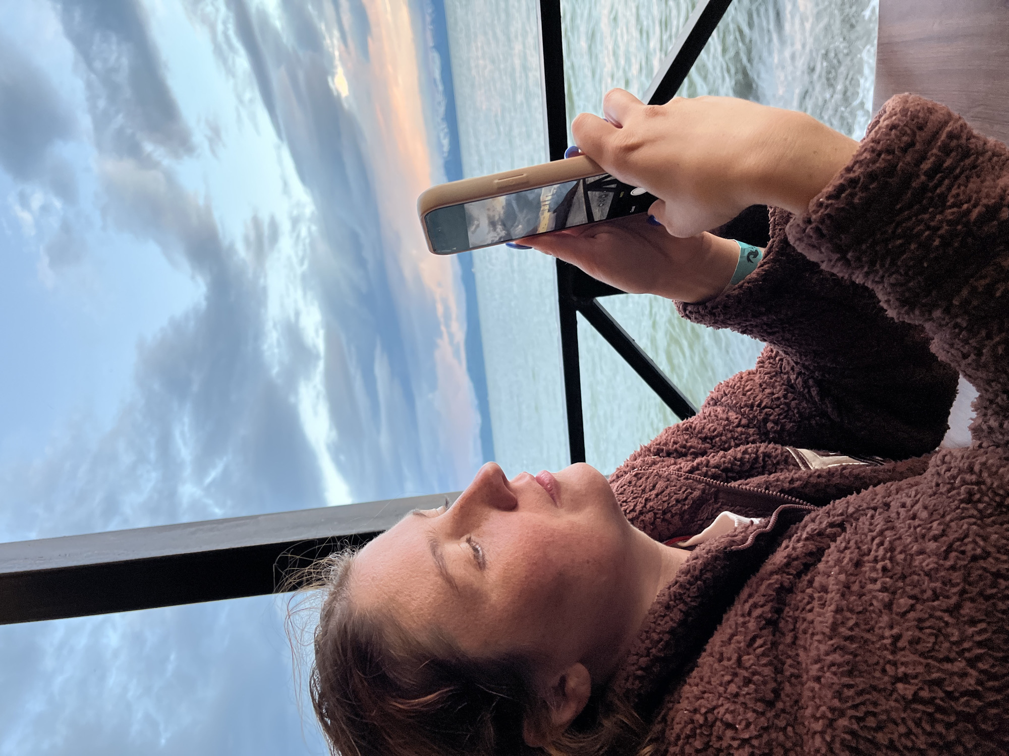A woman with auburn hair, wearing a brown fuzzy jacket, is captured in profile as she holds a smartphone to take a picture. She is seated indoors, with a dark metal railing and a window frame to her left, framing a view of a body of water under a cloudy sky. The water appears to be a sea or large lake, with distant mountains visible on the horizon. The sky is a mix of blues and grays, with hints of pink and orange suggesting either sunrise or sunset. The woman's gaze is directed at her phone, which displays a reflection of the sky and the surrounding environment. The overall setting suggests a serene and contemplative moment by the water. The location is identified as the city of Yelizovo, Russia.