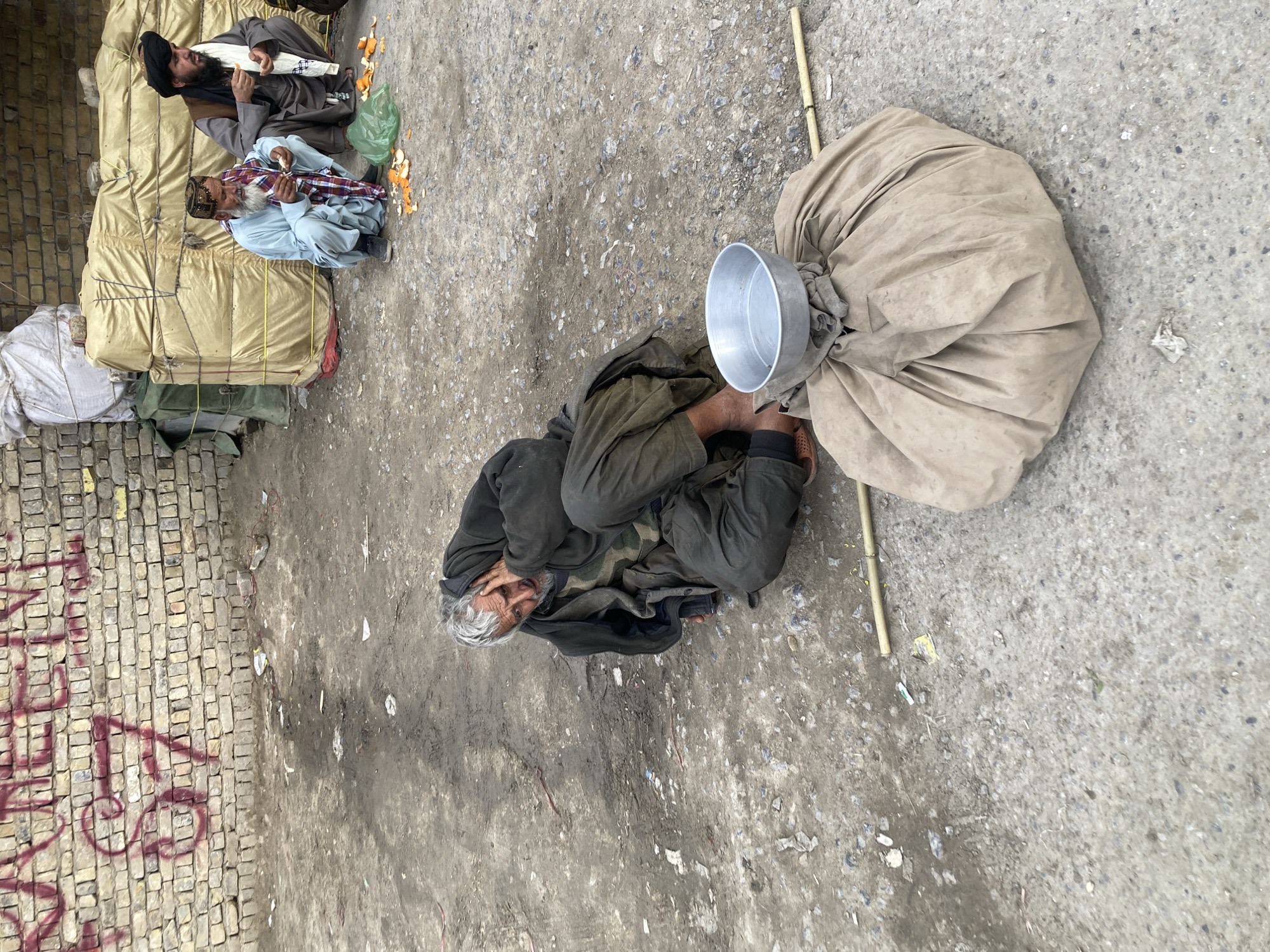 This image depicts an outdoor scene in Quetta City Tehsil, Pakistan. In the foreground, an adult male with grey hair and beard, dressed in dark, layered, and worn clothing, sits on the dirt and gravel ground with his right hand pressed against the side of his head. Adjacent to him, a large, irregularly shaped object wrapped in tan fabric is positioned on the ground, topped with an empty, silver-colored metal bowl. A thin, light-colored wooden stick lies horizontally on the ground to the right of the bundled object.
In the middle ground, two other adult males are observed squatting. The man on the left, with a white beard, wears a light blue shalwar kameez and a patterned cap, actively peeling an orange. The man on the right, with a black beard, wears a dark shalwar kameez and a black turban, holding a piece of orange. Orange peels and a collapsed green plastic bag are present on the ground in front of these two individuals.
The background features large, stacked bundles covered in light-tan tarpaulin, positioned against a light-colored brick wall. Red graffiti is visible on the upper section of the brick wall. The ground across the entire scene is unpaved, consisting of dirt and gravel, with scattered debris and litter.