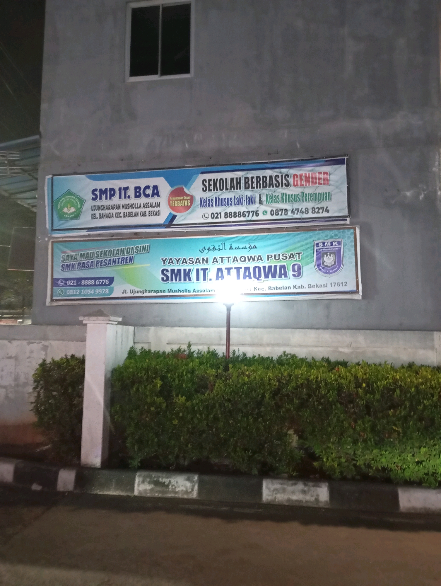 The image shows two banners displayed on the side of a building. The upper banner advertises "SMP IT. BCA" and "SEKOLAH BERBASIS GENDER", with information about accepting students for "Kelas Khusus Laki-laki & Kelas Khusus Perempuan". It also lists contact phone numbers: 021 88886776 and 0878 4748 8274. The lower banner is for "SMK IT. ATTAQWA 9" and states "SAYA MAU SEKOLAH DI SINI SMK RASA PESANTREN". This banner also includes contact numbers: 021-8888 6776 and 0812 1094 9978, and the address "Jl. Ujungharapan Musholla Assalam Kec. Babelan Kab. Bekasi 17612". The banners are mounted on a concrete wall, and below them is a manicured hedge and a paved area. The lighting suggests it is evening or nighttime.