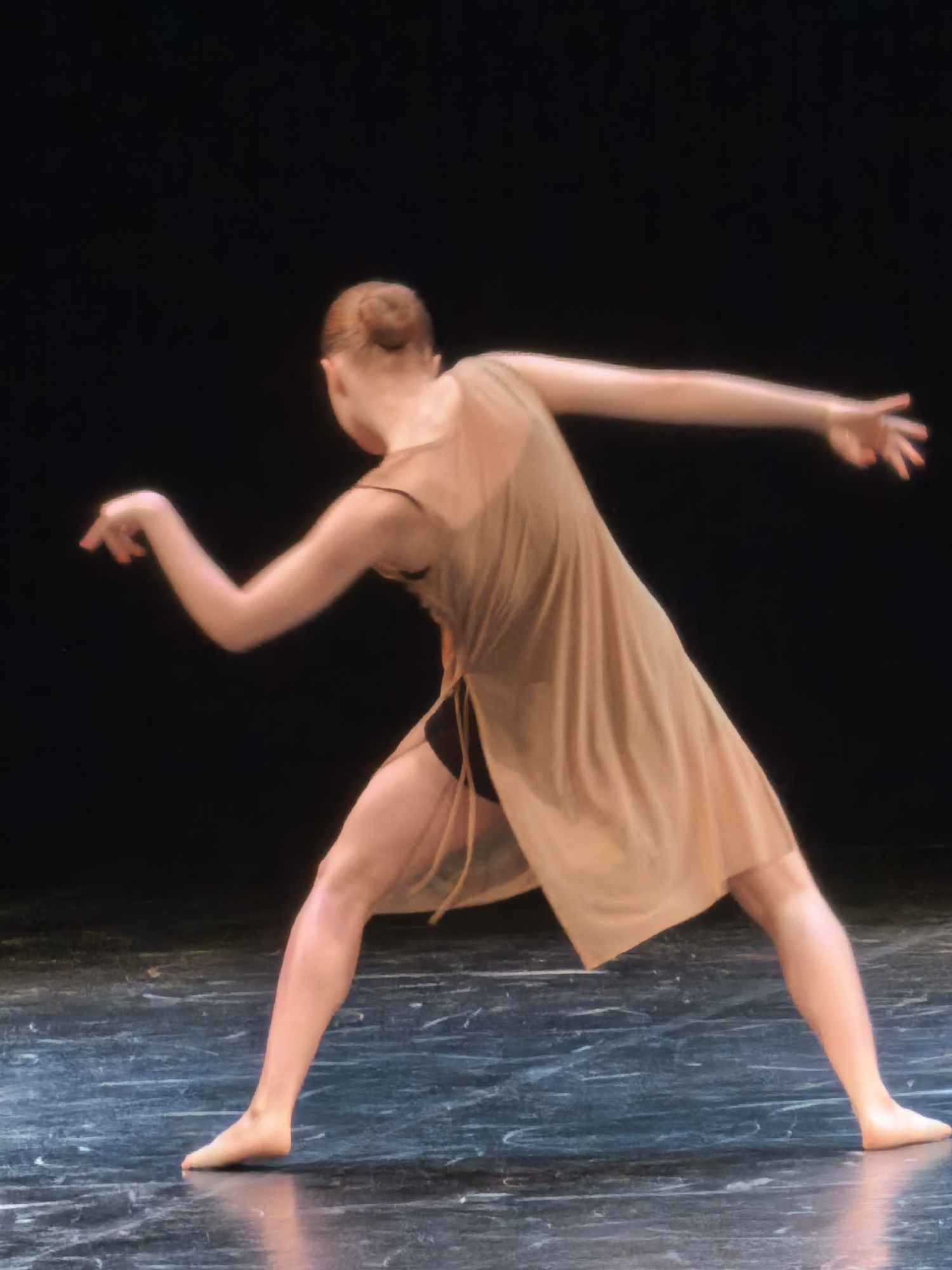 A female dancer with light skin and blonde hair styled in a bun is on a stage with a dark background and a glossy, dark blue floor. She is wearing a sheer, light brown dress over black dance shorts and is in mid-movement, leaning to her left with her left leg extended forward and her right leg bent behind her. Her left arm is extended to the side, and her right arm is bent and held close to her body. Her head is turned to her left, and her expression is not visible. The lighting on the stage is dramatic, highlighting the dancer and casting shadows.