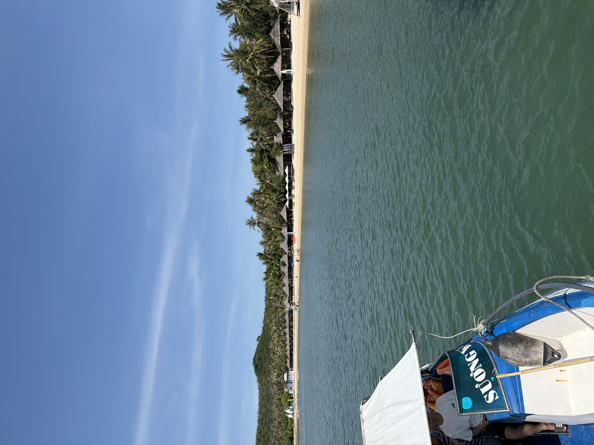 The image depicts a scenic view of a tropical beach in Ninh Ich, Vietnam, viewed from a boat. The foreground shows the bow of a blue and white boat with a white canopy. A person wearing a white t-shirt and black shorts is seated on the boat. The boat has the word "SUONGV" written in white letters on a green panel. The sea in front of the boat is calm and a deep green color.
The middle ground features a sandy beach lined with palm trees. Numerous thatched-roof cabanas and white umbrellas are spread across the beach, suggesting a resort or leisure area. Several people are visible on the beach, some walking, others potentially relaxing under the umbrellas. Surfboards are also seen on the sand.
The background consists of a lush, green, hilly landscape covered in dense vegetation, predominantly palm trees. The sky is clear and blue with a few wispy white clouds. The overall scene conveys a serene and idyllic tropical vacation setting.