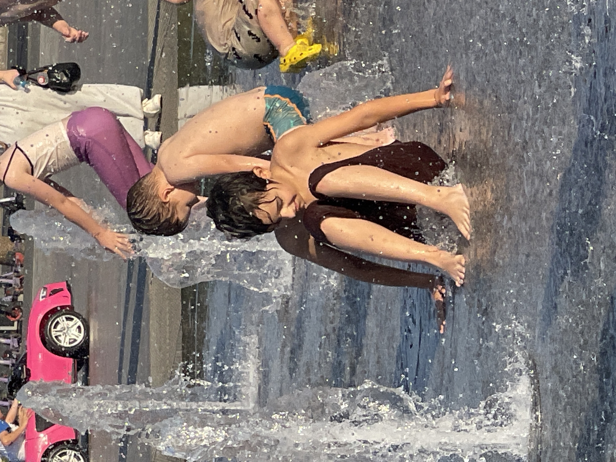 This image depicts an outdoor scene with several children playing in a water fountain. In the foreground, two young boys are seated on the wet pavement, surrounded by splashing water. One boy, wearing dark shorts, is looking down, his hands resting on the ground. The other boy, wearing patterned swim trunks, is partially visible behind him. In the background, a pink toy car is parked, and other individuals, including a person in purple pants and a white top carrying a small black purse, are also present, suggesting a public area or park. The overall environment appears to be a sunny day, with water creating a dynamic and playful atmosphere. The presence of the toy car and the casual attire of the individuals suggest a leisure activity, possibly in a city park or public square. The scene is set in Orenburg, Russia.