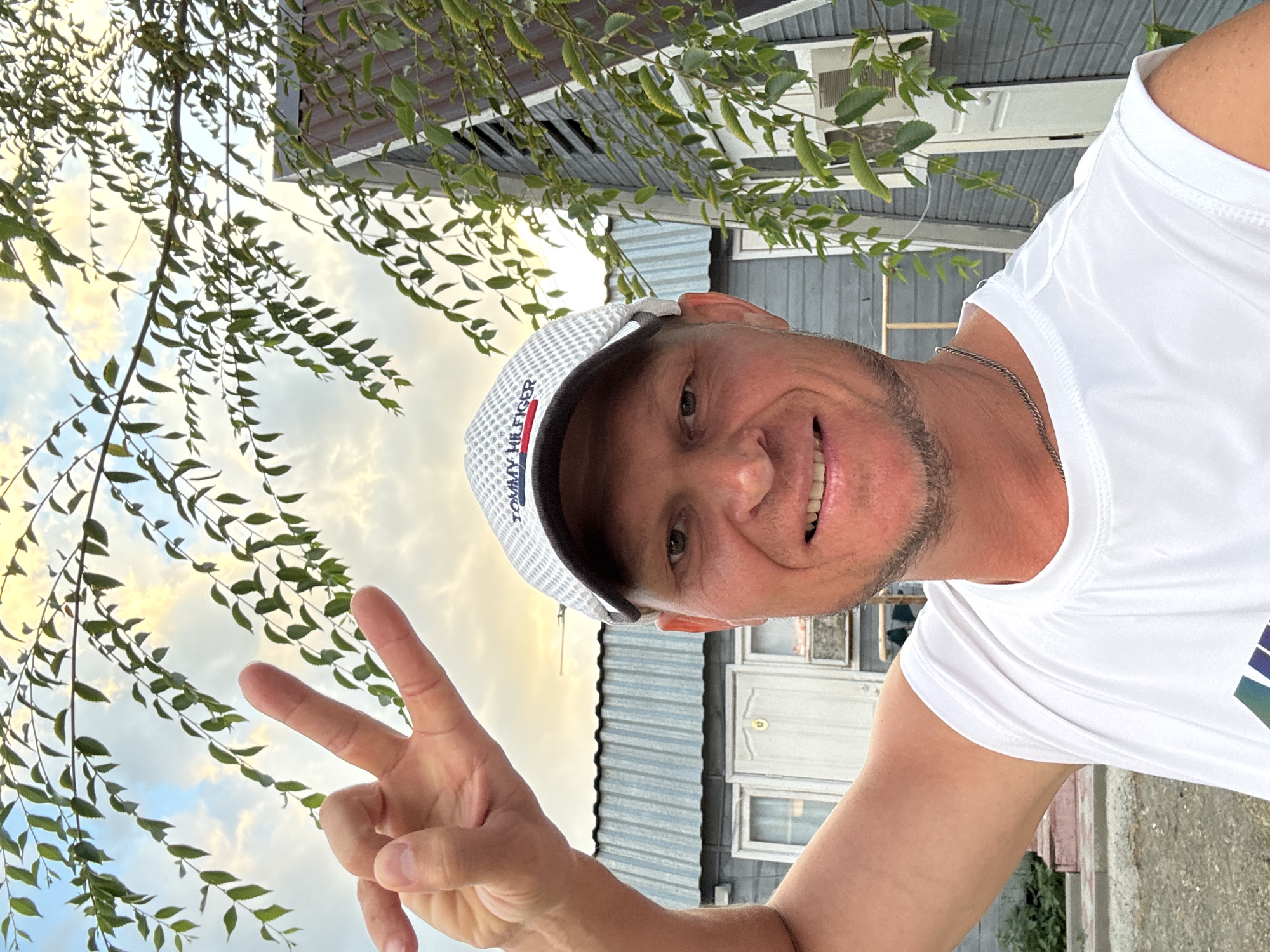 A man with fair skin, short-cropped blonde hair, and a light beard is taking a selfie outdoors. He is wearing a white t-shirt and a white baseball cap with the "Tommy Hilfiger" logo. He is smiling and making a peace sign with his right hand. Behind him is a grey wooden building with a dark metal roof. There are also some green branches with leaves hanging in the foreground, partially obscuring the sky. The sky appears to be cloudy with some yellow and orange hues, suggesting it might be sunset or sunrise. The ground in front of the building is rough and appears to be made of dirt or gravel. The building has windows and a door with the number "12" on it. An air conditioning unit is visible on the exterior wall of the building. The location is the city of Zharbulak, Kazakhstan.