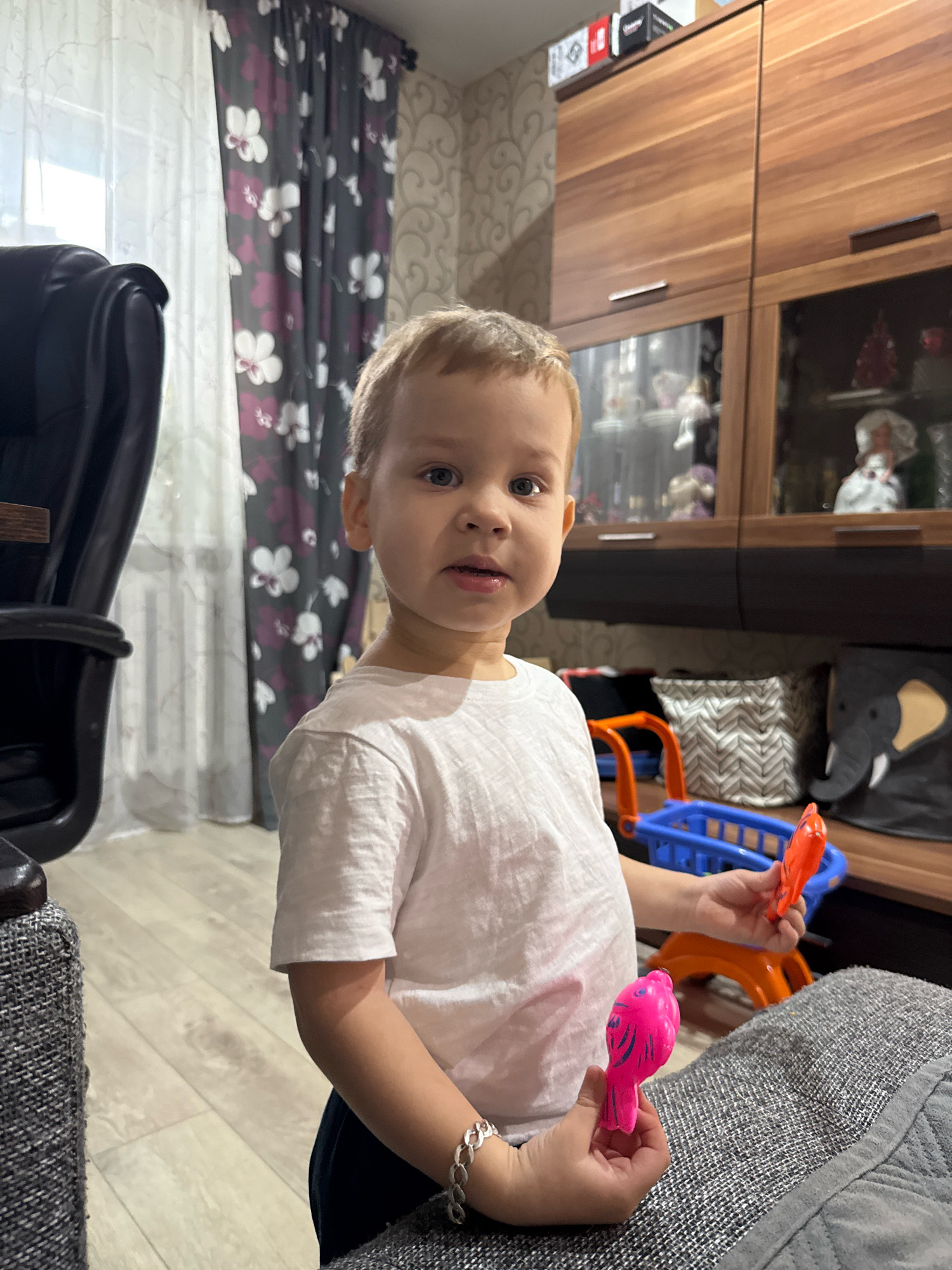 A toddler with blonde hair and blue eyes, wearing a white t-shirt and dark pants, stands in a living room. The child holds a pink toy bird in their left hand and an orange toy in their right hand. In the background, a blue and orange toy shopping cart is visible. The room features light wood flooring, floral curtains, and a wooden cabinet filled with decorative items. A grey upholstered chair and a dark leather chair are also present. The image does not provide any specific details indicating the location as Rusanovo, Russia.