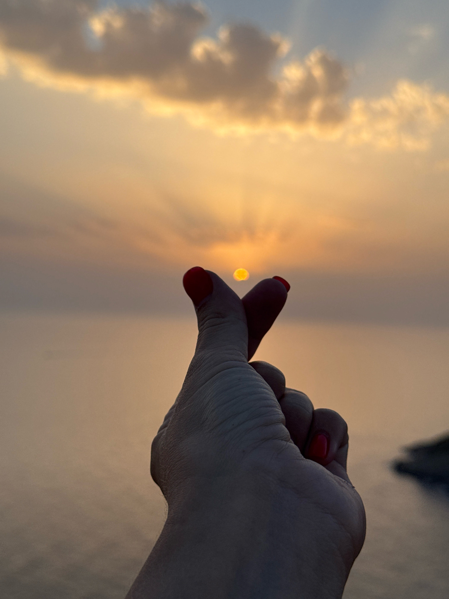 A human hand, with bright red nail polish on the thumb and index finger, is held up in the foreground against a hazy sunset sky over a body of water. The thumb and index finger are pressed together to form a heart shape, with the sun appearing to be contained within the shape. The sky is a gradient of orange, yellow, and pale blue, with wispy clouds. The water is a muted orange-brown. A faint outline of land is visible on the right side of the image. The image is a photograph taken at dusk. The location is identified as Zagora, Montenegro.