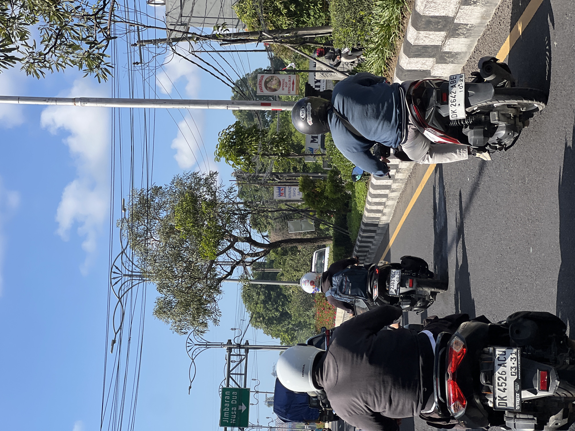 The image captures a bright, sunny day in Kuta, Indonesia, with a clear blue sky and scattered white clouds. The scene focuses on a street with several motorbikes in motion. In the foreground, a person wearing a white helmet and a dark jacket rides a black motorbike. Behind them, another individual in a white helmet and a dark backpack is on a black motorbike, followed by a third person wearing a black helmet and a blue jacket, also on a motorbike.
To the left of the road, a green sign indicates directions to "Jimbaran Nusa Dua." Trees and vegetation line the roadside, with some buildings and overhead power lines visible in the background. The road itself has a yellow lane marker and is bordered by a concrete barrier with horizontal stripes. The overall impression is of a typical day on a busy street in Kuta.
