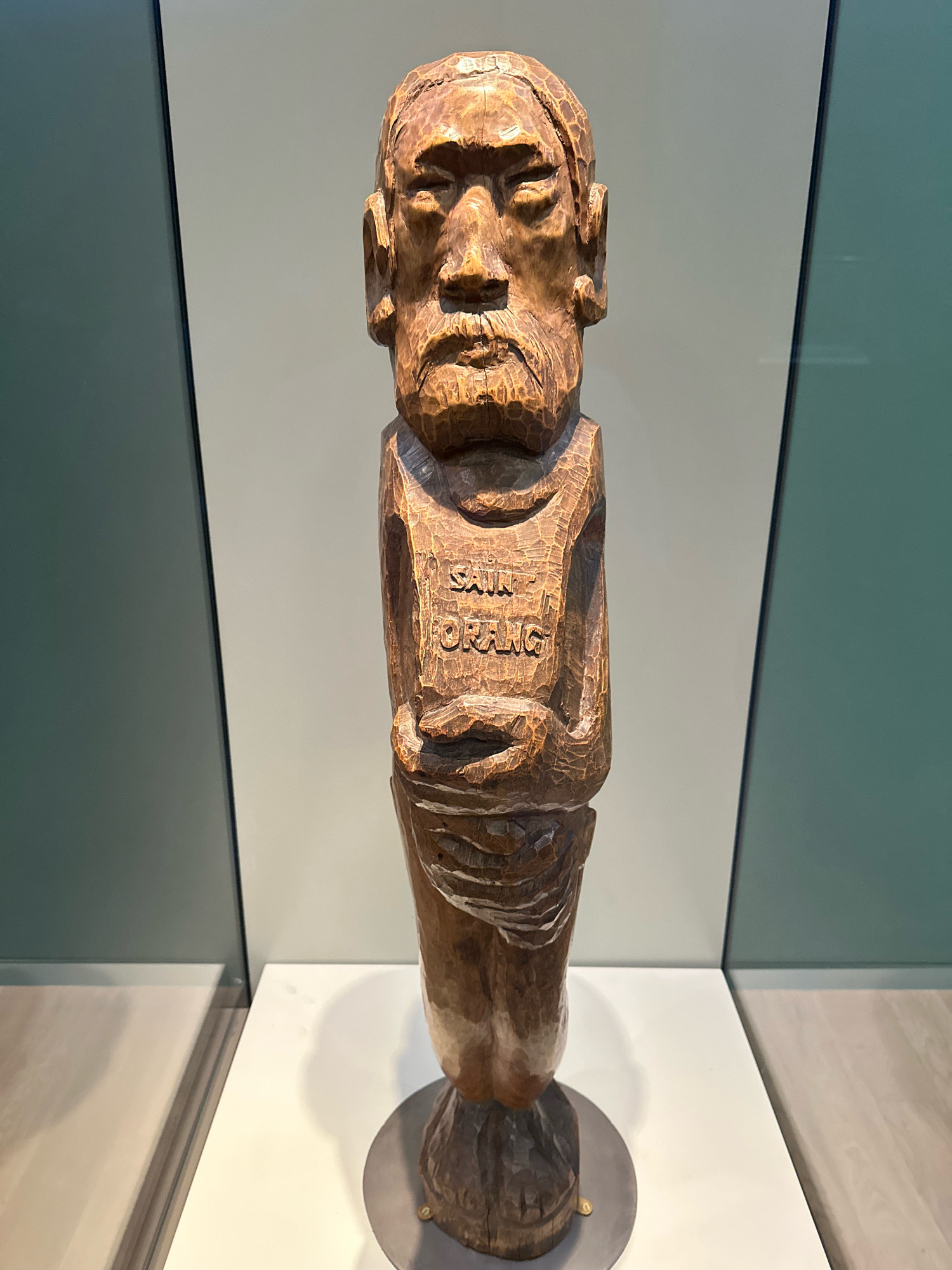 A full-shot, front-facing wooden sculpture of a man stands on a white pedestal in a museum exhibit. The sculpture depicts a man with distinct facial features, including carved eyes, a nose, and a beard. His body is clad in what appears to be a garment with the words "SAIR ORANG" carved onto the chest. The sculpture is made of dark wood and shows signs of wear and age, with visible grain and tool marks. The background consists of two vertical panels, one dark gray on the left and one light gray on the right, separated by a thin black line. The exhibit space is well-lit, with subtle reflections visible on the floor in the foreground. The sculpture is centrally positioned and the focus of the image.