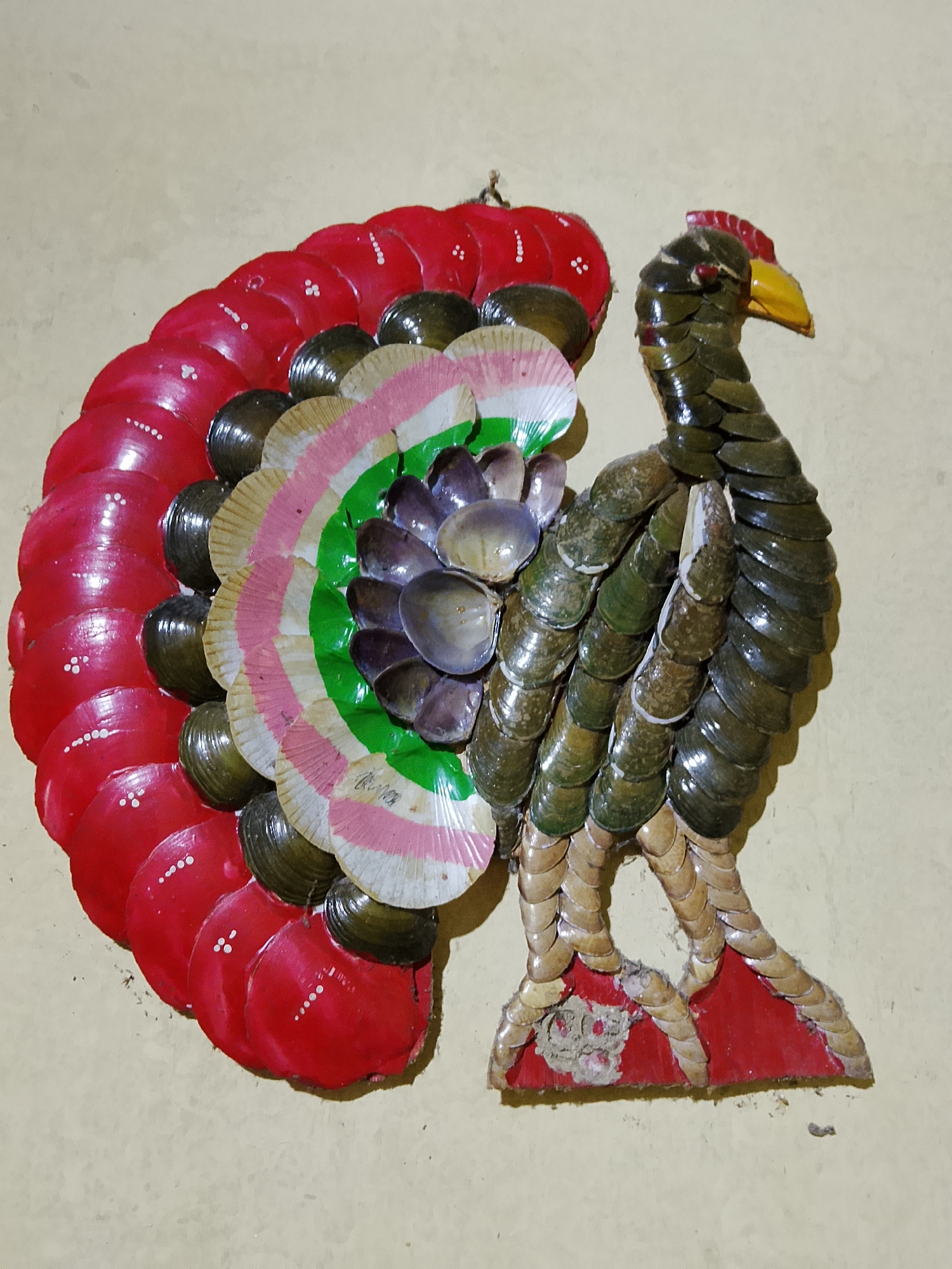 This is a close-up, eye-level, medium shot of a decorative wall hanging of a peacock, likely made from seashells and other natural materials, affixed to a textured, light-colored wall. The peacock is depicted facing right, with its tail fanned out. The tail feathers are arranged in a semi-circular pattern, with the outer feathers painted bright red and decorated with small white dots. Moving inward, the feathers transition to dark green, then a gradient of light tan and white with pink stripes, and finally a cluster of darker, iridescent shells. The peacock's body is constructed from overlapping dark green shells, forming its neck, head, and torso. It has a yellow beak and a red crest. Its legs are made of light-colored, stacked shells, and its feet are attached to a red base, which appears to be wooden and has some wear and tear. The background wall is a uniform, light beige or cream color with a slightly rough texture. The lighting appears to be natural or soft artificial light, casting subtle shadows on the wall. The image does not provide any specific identifiable landmarks of Kasiman, Indonesia, beyond the context provided in the prompt.