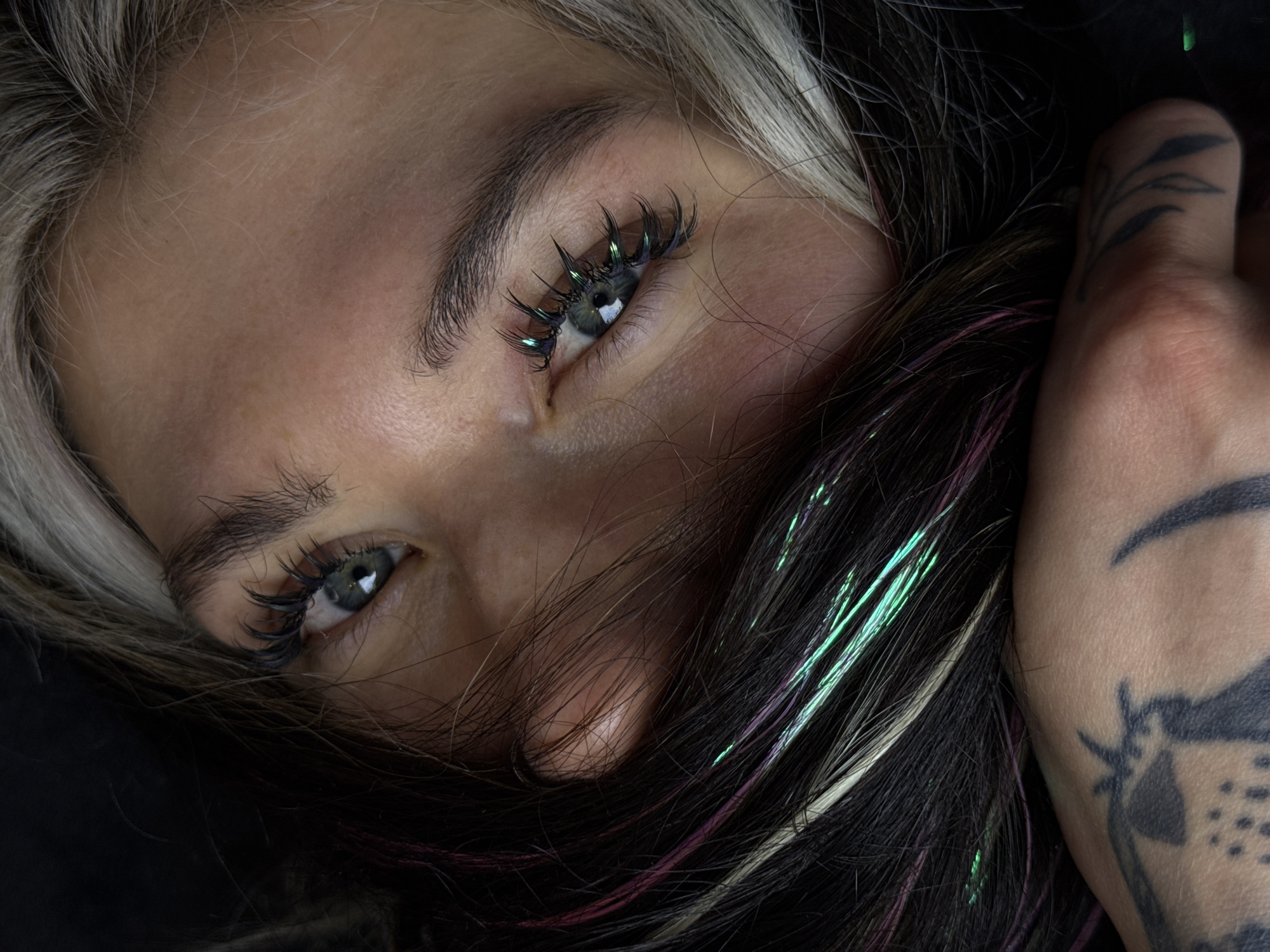 A close-up shot of a person's face, focusing on their eyes. The individual has light-colored skin and grey eyes. The person's hair is a mix of blonde and dark strands, with iridescent strands. The person has long eyelashes. A tattooed hand is visible, grasping the hair.
The location is not provided, and it is not possible to determine the city or environment from the details provided.