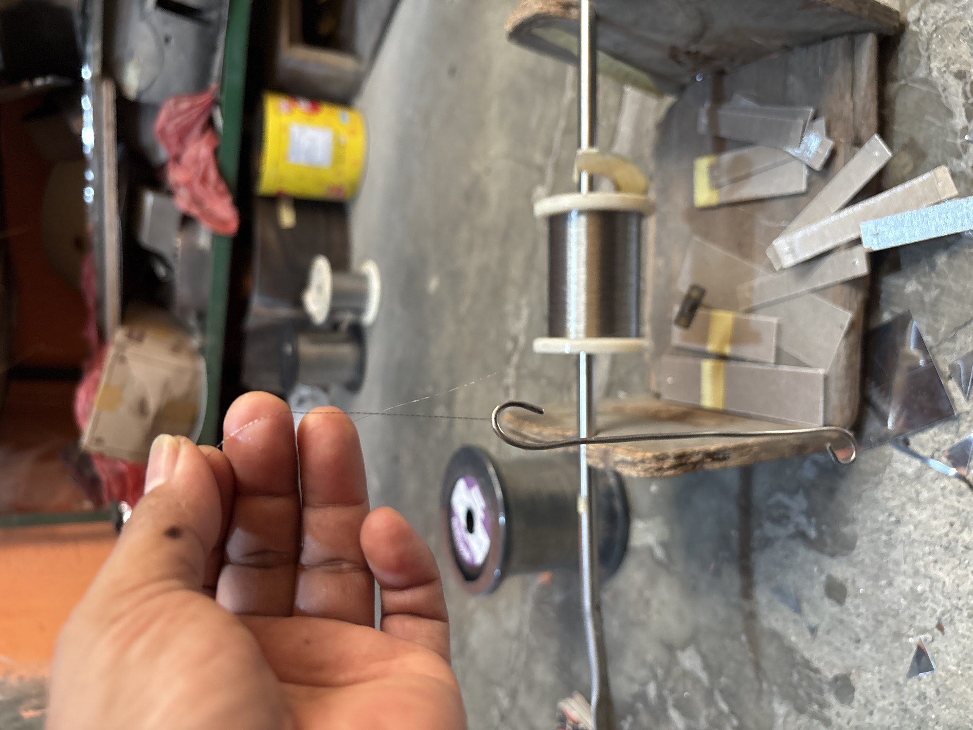 A hand is holding a thin strand of metallic wire, which is attached to a fishing hook. The wire is draped over a metal rod supported by a wooden apparatus. In the background, a spool of fishing line and various other spools and containers are visible, suggesting a workshop or storage area. The overall scene appears to be outdoors or in an open-air setting, with a concrete ground surface and a brick wall in the distance. The lighting suggests it is daytime.