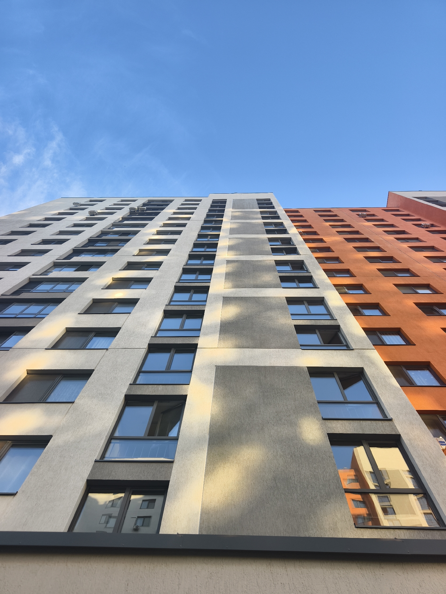 The image shows a low-angle view of a tall building in Tyumen, Russia, against a blue sky with sparse clouds. The building has a modern design with a white and gray facade on one side and an orange facade on the other. Numerous rectangular windows are visible across the building's surface. Some windows reflect the surrounding buildings, particularly the orange facade. Air conditioning units are attached to some of the building's exterior walls. The base of the building is a light beige color.