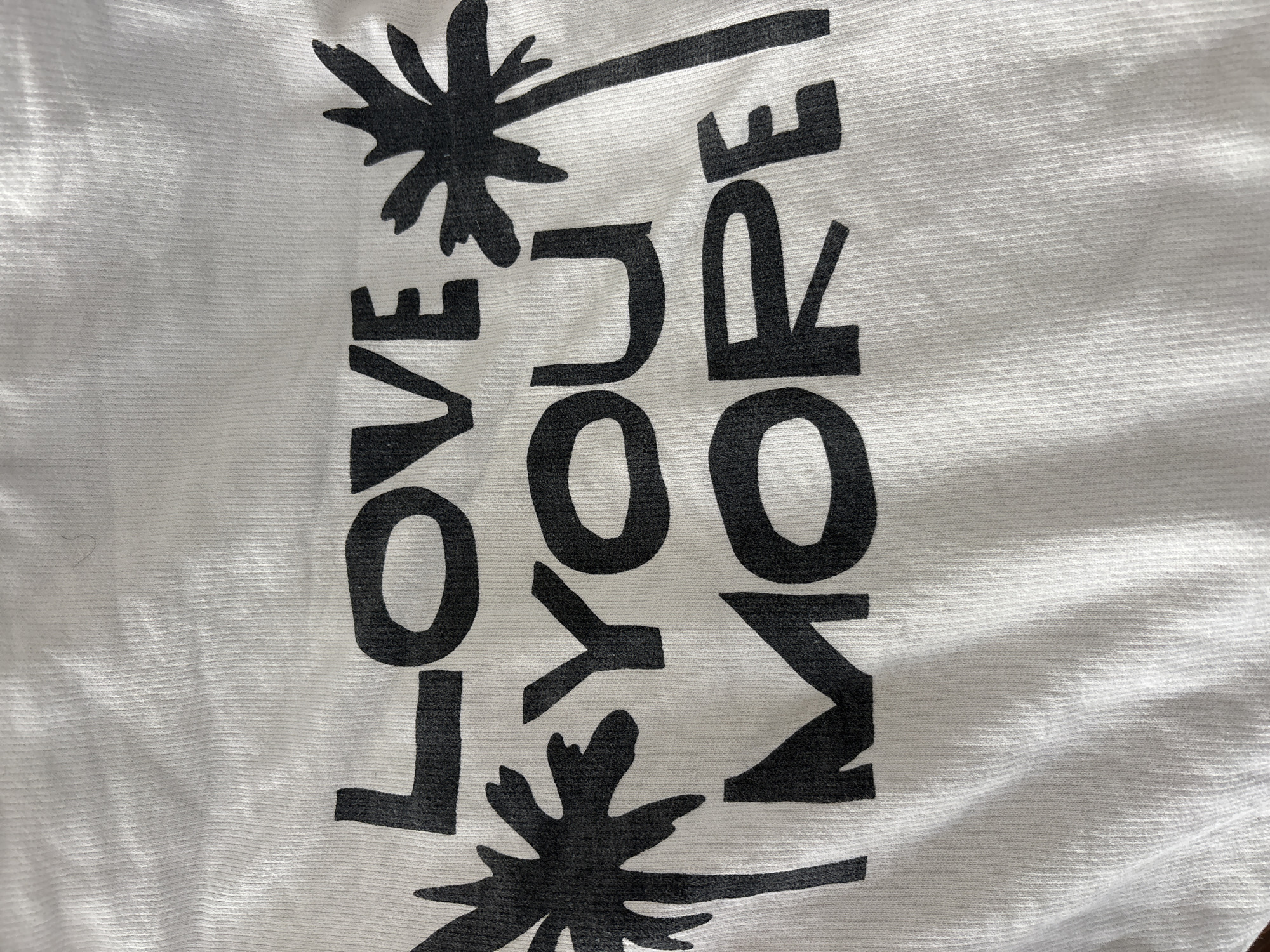 A close-up photograph shows a white fabric, possibly a t-shirt, with black block letters and palm tree designs printed vertically. The text reads "LOVE YOU MORE" with palm trees above and below the words. The image provides no information about the location in Seal Beach, USA or any subjects, objects, actions, or interactions beyond the text and design printed on the fabric.
