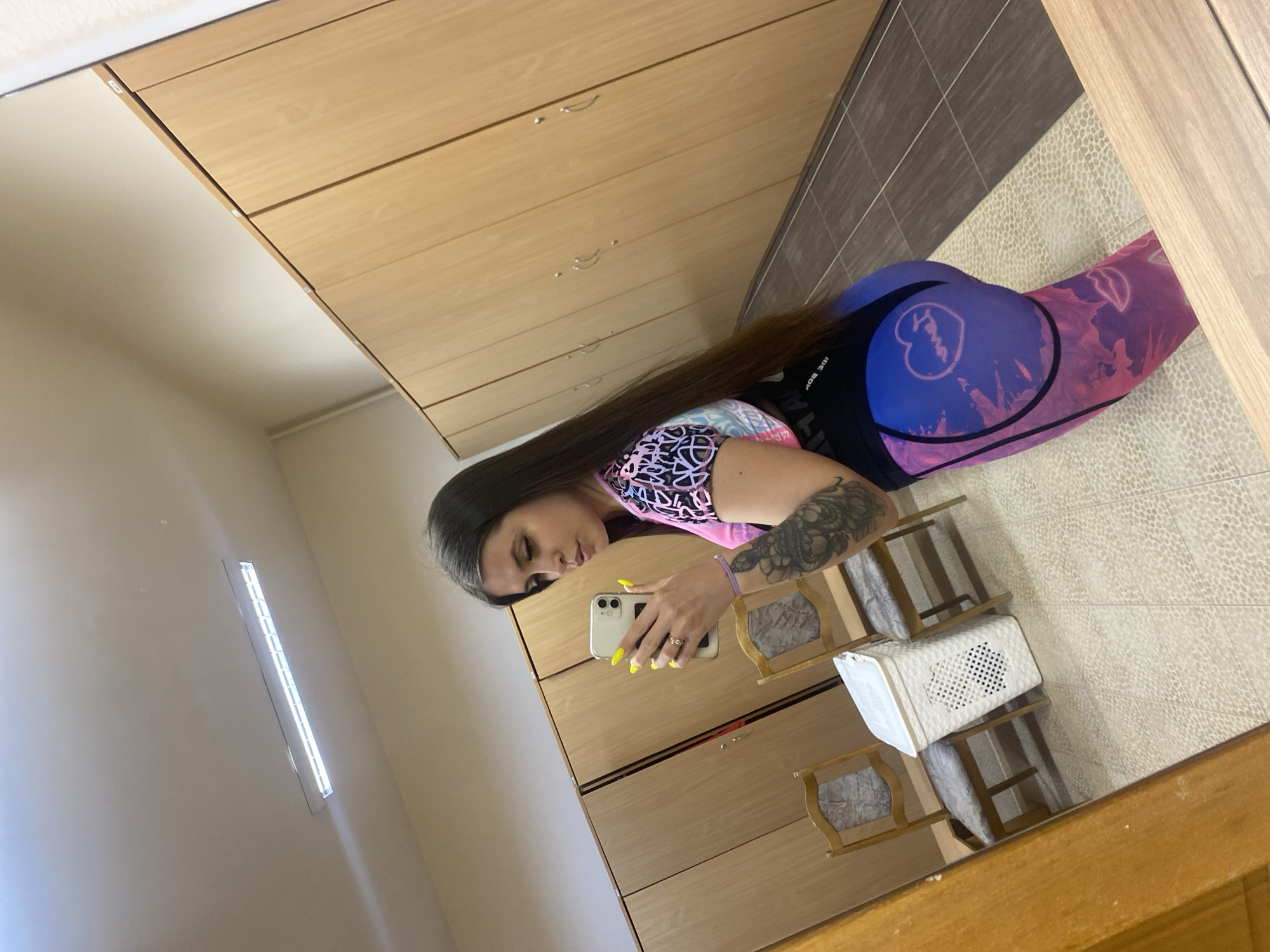 A young woman takes a selfie in a mirror in Chaikovsky, Russia. She holds a smartphone in her right hand and wears leggings with a blue and pink tie-dye pattern. Her dark hair is long and straight. A large tattoo covers her left forearm. The background features a light wood cabinet with multiple doors or drawers, as well as a tiled floor and a white storage basket on a stand. A long, thin light fixture is visible on the wall in the background.