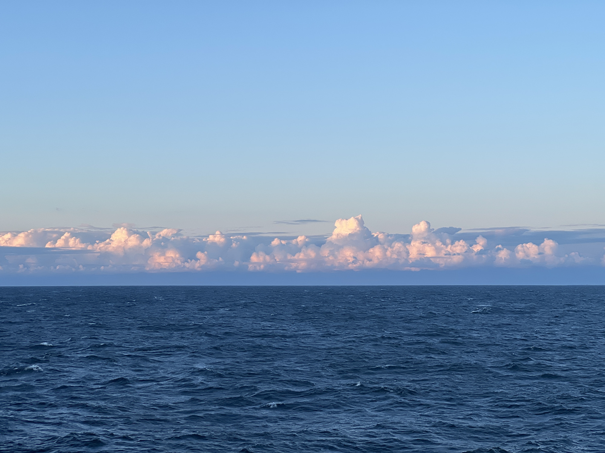 An expansive view of the Baltic Sea under a clear, light blue sky. A line of fluffy, pink-tinged clouds stretches across the horizon, illuminated by the setting or rising sun. The deep blue water is choppy, with small white waves cresting on its surface. The scene is devoid of any human presence or man-made structures, creating a sense of vastness and solitude. The location is unknown, but given the visual context, it is likely a coastal area of Poland.