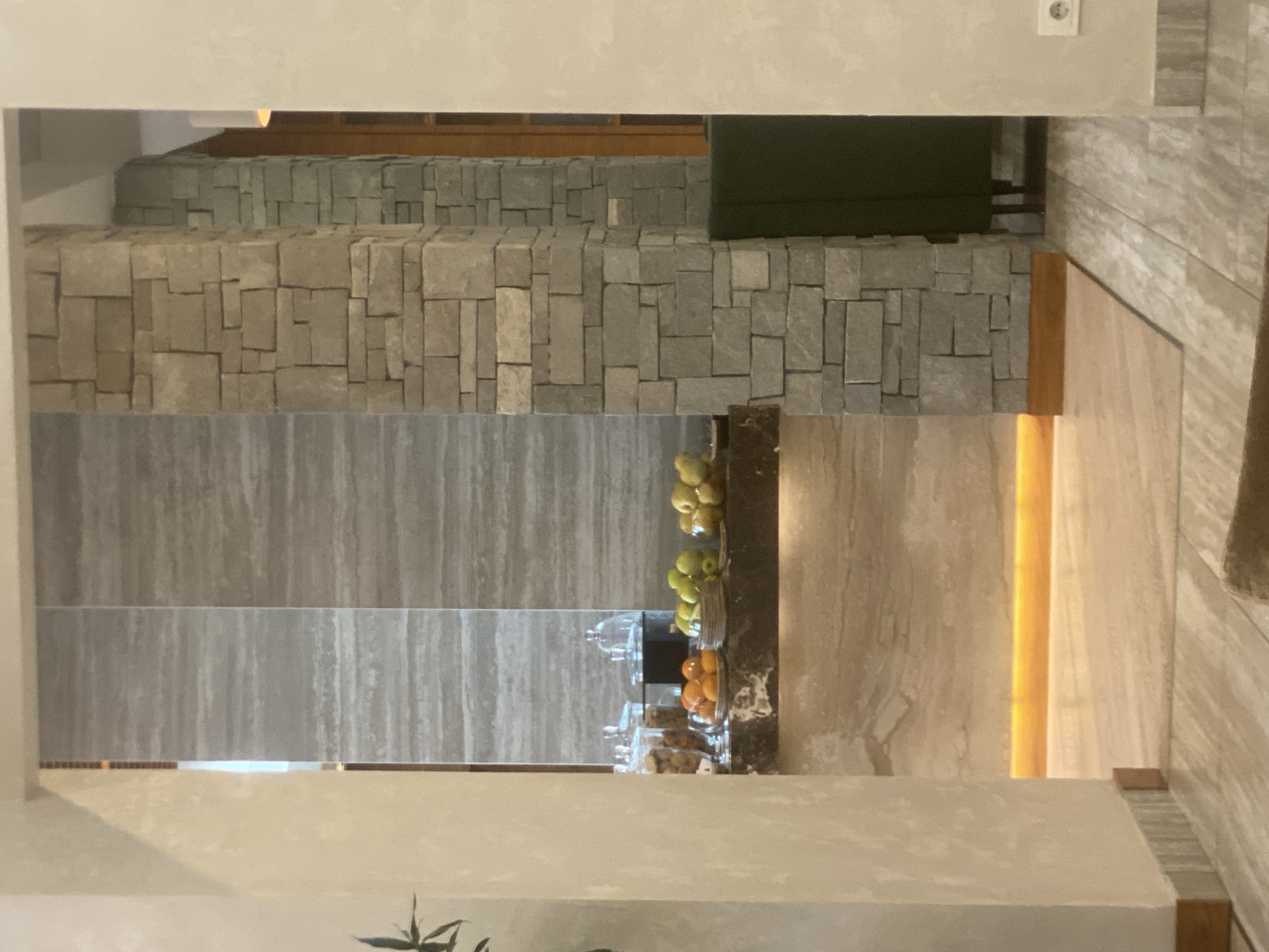 This image captures an interior scene, likely a hotel lobby or a cafe, with elements of natural stone and wood. In the foreground, a light-colored carpet is visible on a tiled floor. To the left, a textured, light-colored wall frames the view into the space. The central area features a counter or bar area with a marble-like countertop. On the counter, several glass jars filled with cookies and bowls of fruit, including oranges and apples, are arranged. Behind the counter, vertical panels with a striated gray pattern are visible. To the right of the counter, a substantial stone pillar constructed from irregularly shaped grey stones stands prominently. Further to the right, a glimpse of dark green upholstery and wooden structures suggest seating or architectural details. An electrical outlet is visible on a light-colored wall in the far right. The scene is illuminated by warm, ambient lighting, particularly a strip of orange light running along the base of the counter area, creating a soft glow. There are no people visible in the image. The overall impression is one of modern, naturalistic interior design.