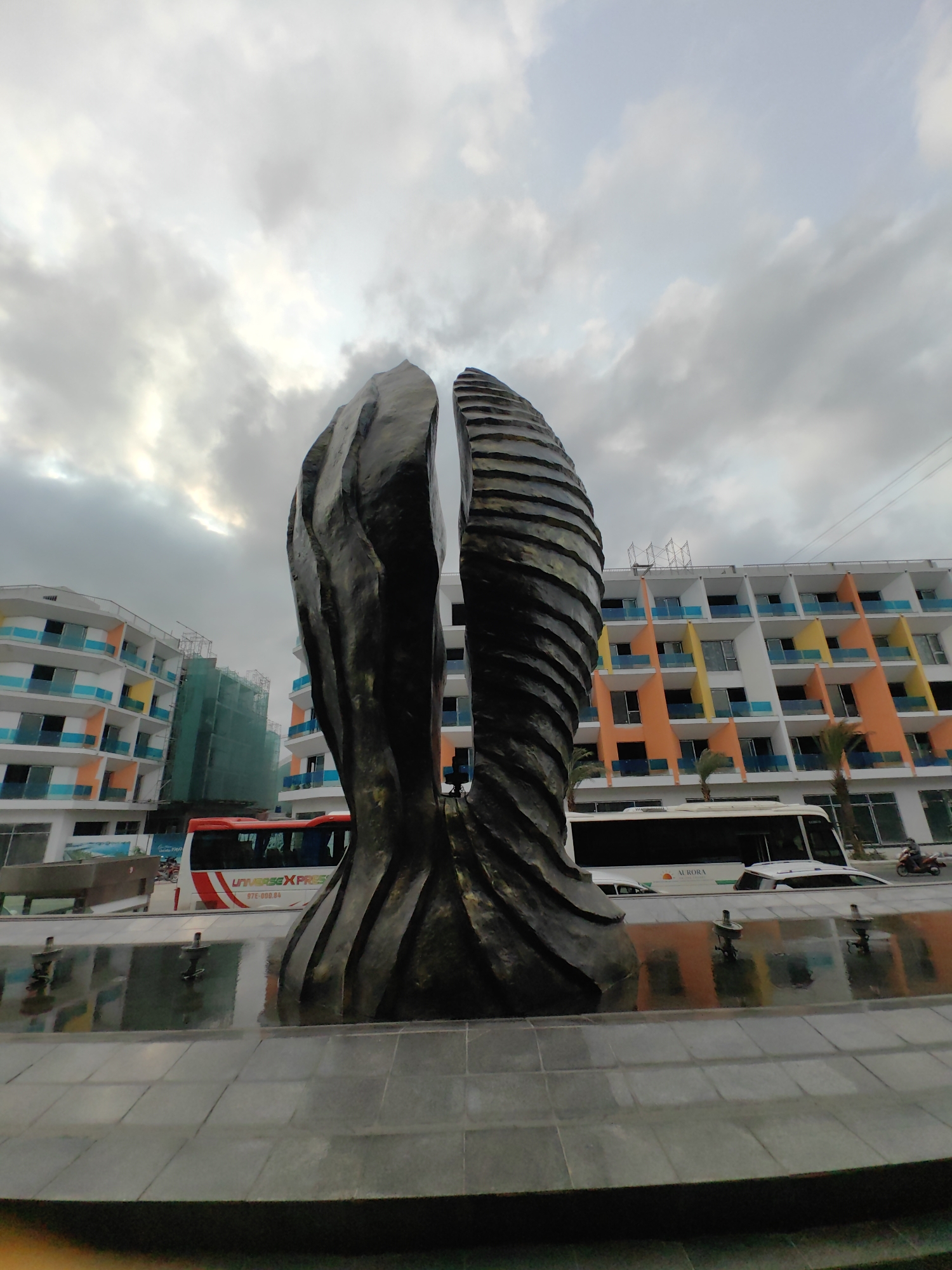 The image presents an urban scene in Bai Chay Ward, Vietnam, dominated by a large, abstract sculpture in the foreground. The sculpture, crafted from a dark, textured material, comprises two distinct, vertical forms. The left form features flowing, undulating lines, while the right form has prominent, horizontal, layered ridges. These forms rise from a shallow water feature, which subtly reflects the surrounding environment.
In the mid-ground, vibrant multi-story buildings line the street. These structures feature numerous balconies with striking blue, orange, and yellow panels, giving them a modern and colorful aesthetic. One building on the left is partially covered by green construction scaffolding, indicating ongoing development.
On the road, several vehicles are visible. A red and white bus, bearing the text "UNIVERSE XPRESS" and the license plate "97E-000.84", is parked near the left building. Another white bus with "AURORA" written on its side is seen further to the right. A white car and a red scooter are also present.
The sky is overcast with large, diffused grey clouds, suggesting either an early morning, late afternoon, or simply a cloudy day. No people are prominently visible, contributing to a tranquil, almost static atmosphere despite the urban setting. The scene highlights contemporary architecture and public art within a developing area.