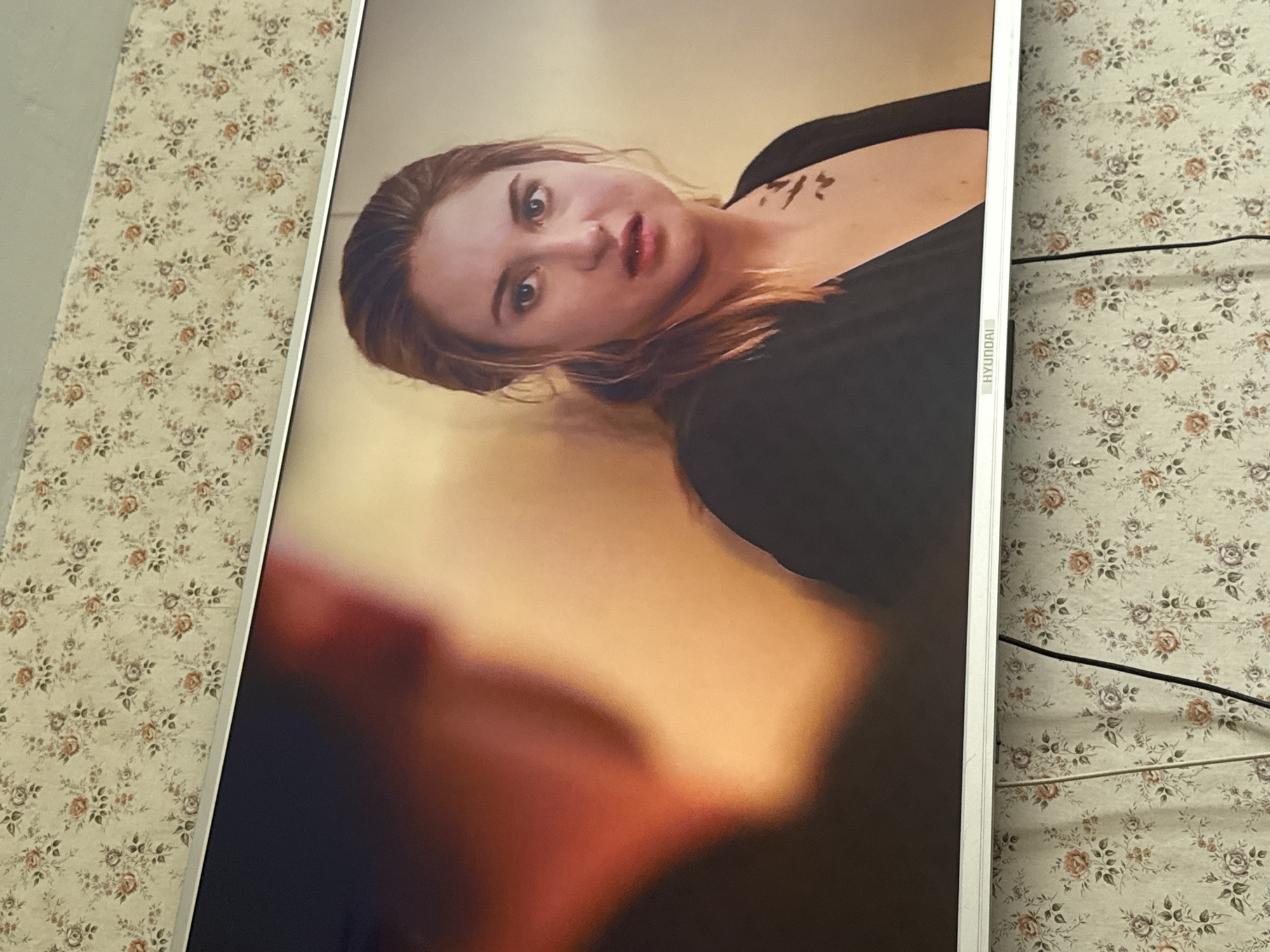 The image shows a Hyundai TV screen displaying a woman in a black top with a tattoo on her chest, looking towards the viewer. The screen is placed against a background of floral patterned wallpaper. Electrical wires are visible on the right side. The image was taken indoors, presumably in a residential setting. No information is provided to confirm its location in Mytishchi, Russia.