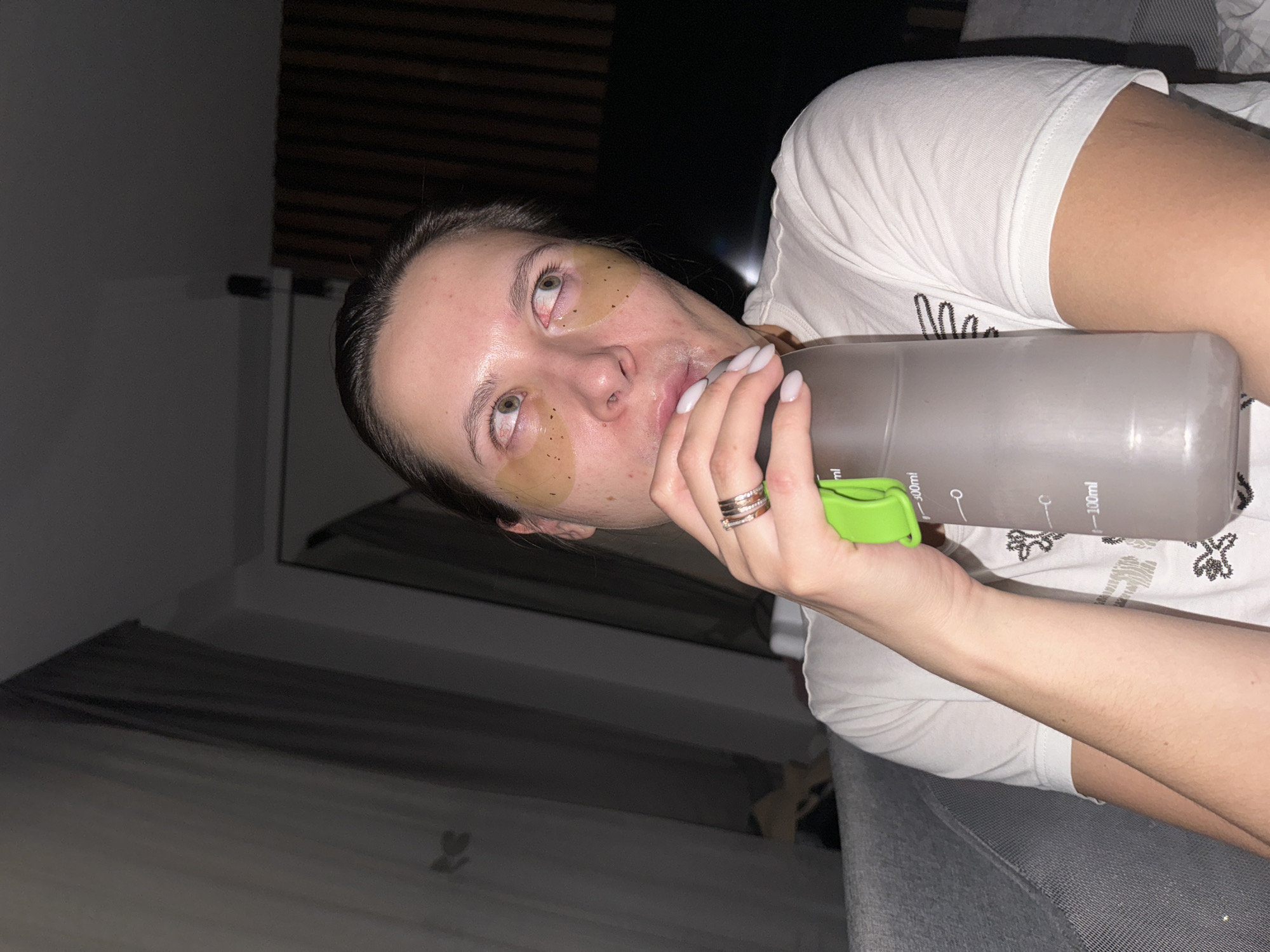 A woman with under-eye patches is holding a water bottle with a green lid in what appears to be an indoor setting. The woman is wearing a white t-shirt and multiple rings on her finger. The background shows a wall and the edge of a sofa or bed. There is insufficient information to determine if the location is in Akulovo (Gp Kubinka), Russia.