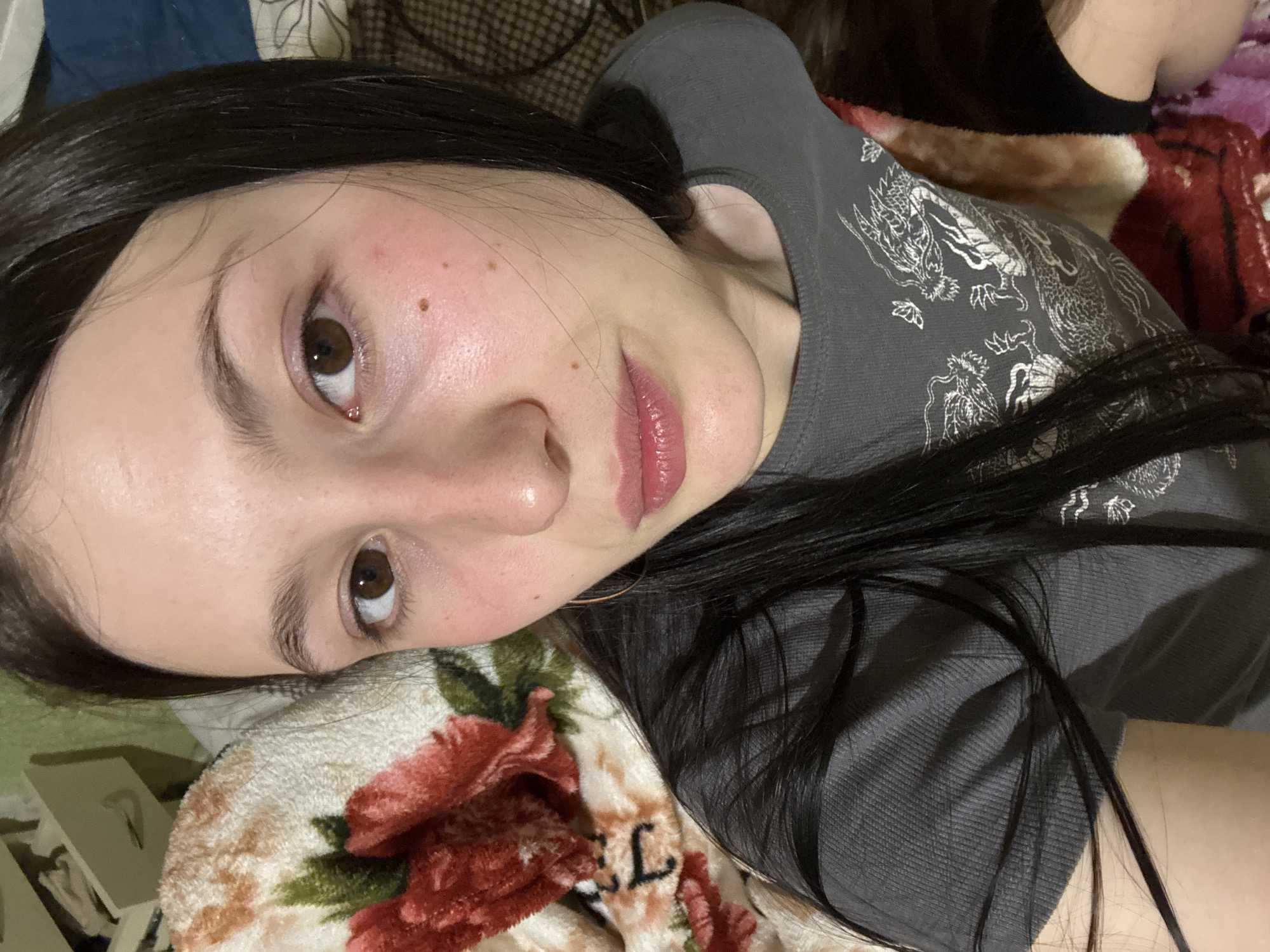 A young woman with fair skin and dark hair is lying down. She has brown eyes, a mole on her cheek, and pink lips. She is wearing a grey t-shirt with a dragon design. Her head rests on a floral-patterned pillow. The background includes a blue fabric and a plaid pattern. There is no evidence in the image to suggest the location is Фоки, Russia.