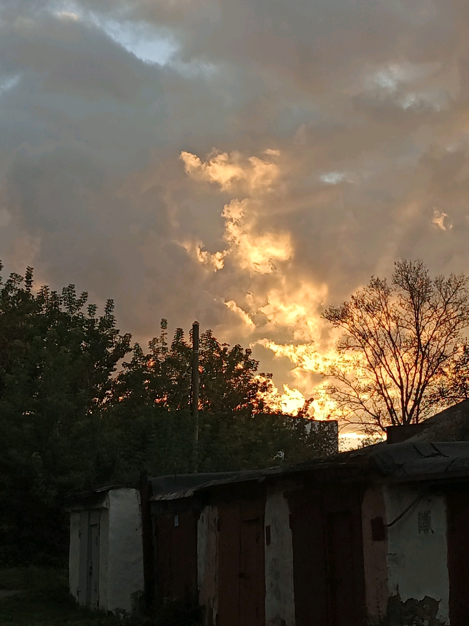 This image captures a dramatic sunset over Zlatoust, Russia. The sky is filled with turbulent clouds, illuminated by the setting sun with hues of orange and yellow. Silhouetted against this vibrant sky are dark green trees and a bare tree with intricate branches on the right. In the foreground, a row of old, weathered garages or storage units runs horizontally across the bottom of the frame. These structures appear to be made of concrete and metal, with visible signs of age and disrepair, including peeling paint and rusted metal roofs. The overall impression is one of a natural spectacle unfolding above a mundane, industrial or utilitarian setting.