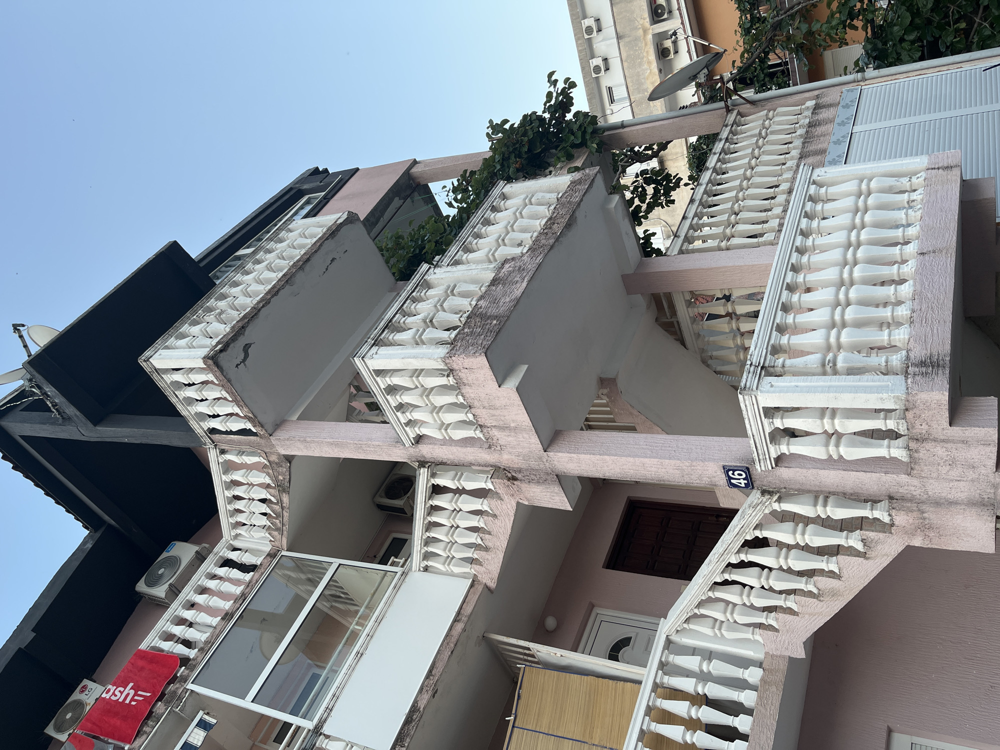 An outdoor, low-angle, full shot shows the exterior of a pink, multi-story apartment building located at address number 46 in the city of Budva, Montenegro. The building has a dark, angled roof. White balustrades with ornate posts line several balconies and an outdoor staircase. The staircase, which is part of the building's exterior, is also pink and has white balustrades. An air conditioning unit is mounted on the wall of the building. A red towel hangs from a line on the upper left side of the building. The sky is clear and light blue.