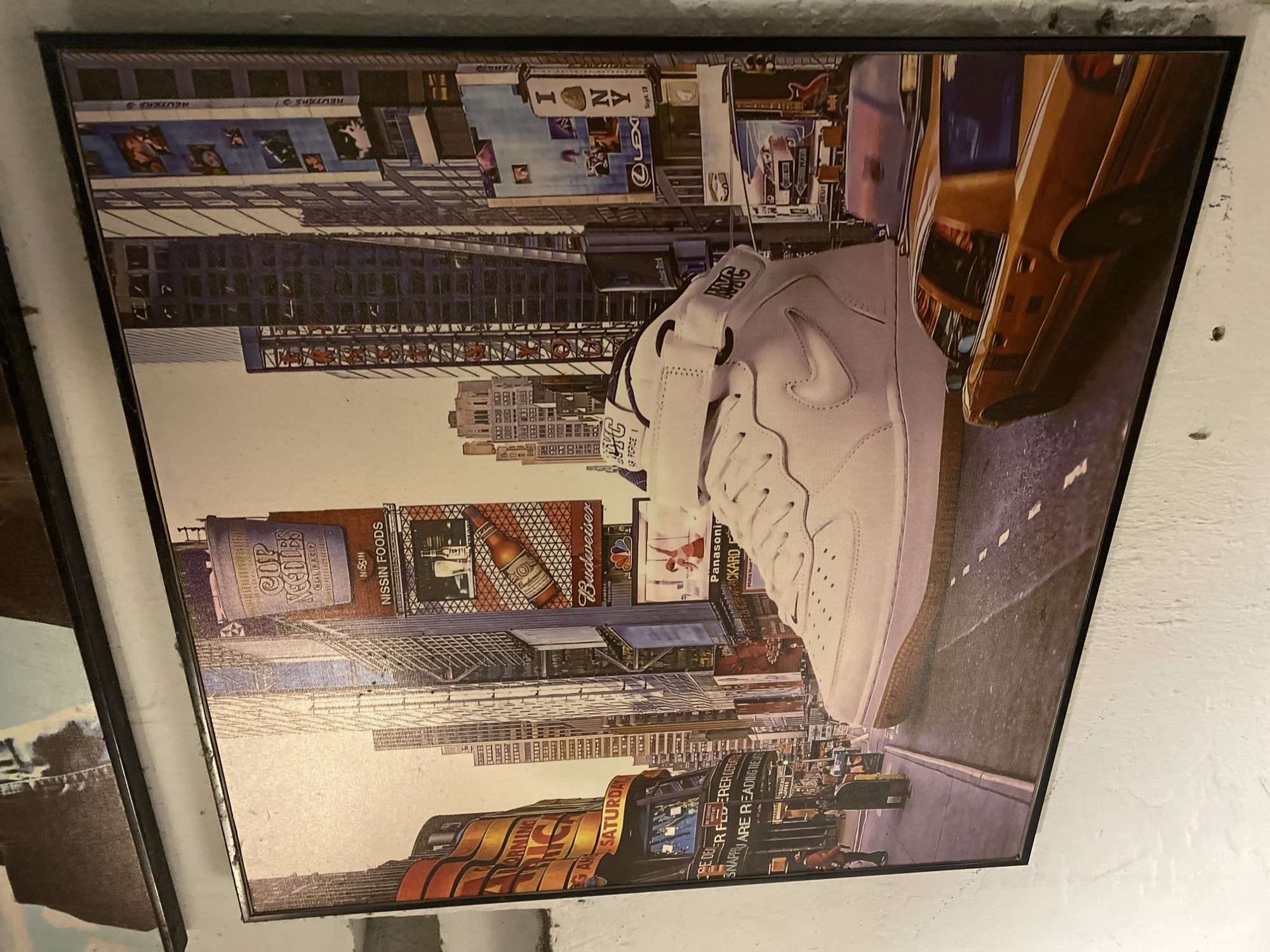 This is a vertically oriented, framed print depicting a collage of images, with the most prominent subject being a white Nike Air Force 1 sneaker positioned centrally. The sneaker is viewed from the side, with the laces and part of the sole visible.
The background is a busy, urban scene reminiscent of Times Square, New York City. It features numerous illuminated billboards and building facades, showcasing various advertisements and text. Some identifiable text includes "I NY," "Budweiser," "NISSIN FOODS," "CUP NOODLES," "Panasoni CKARD," and "LEX." The overall atmosphere suggests a nighttime or twilight setting due to the illuminated signs. A yellow taxi is visible on the right side of the image, further reinforcing the New York City context. The style of the collage suggests a focus on urban culture and iconic branding. The print appears to be mounted on a textured white wall, with the frame being black.