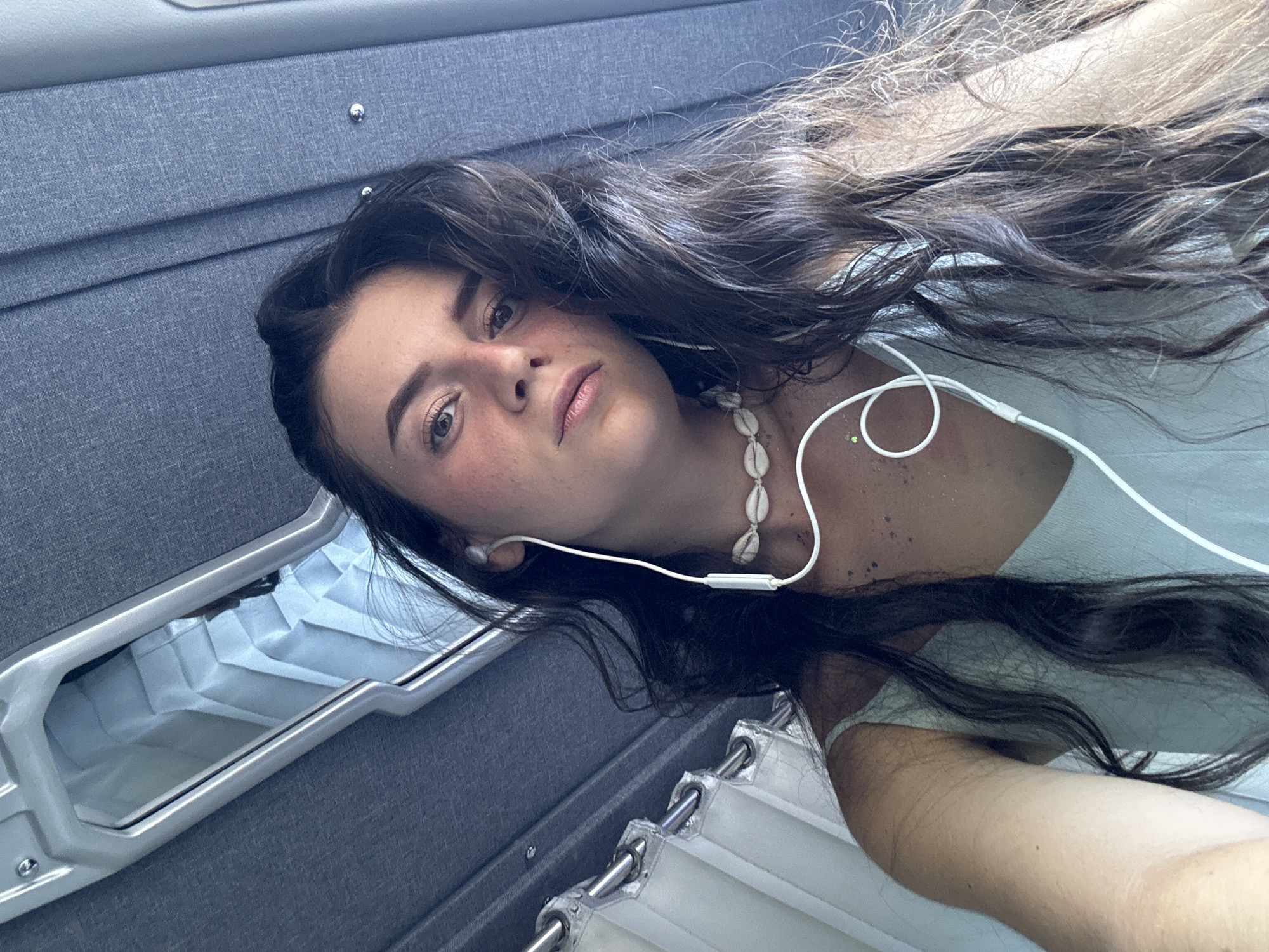 A young woman with long, dark, wavy hair lies on a gray upholstered surface with a metal handle and a white curtain. She is wearing a light-colored top, white earphones, and a seashell necklace. The location cannot be determined from the provided image.