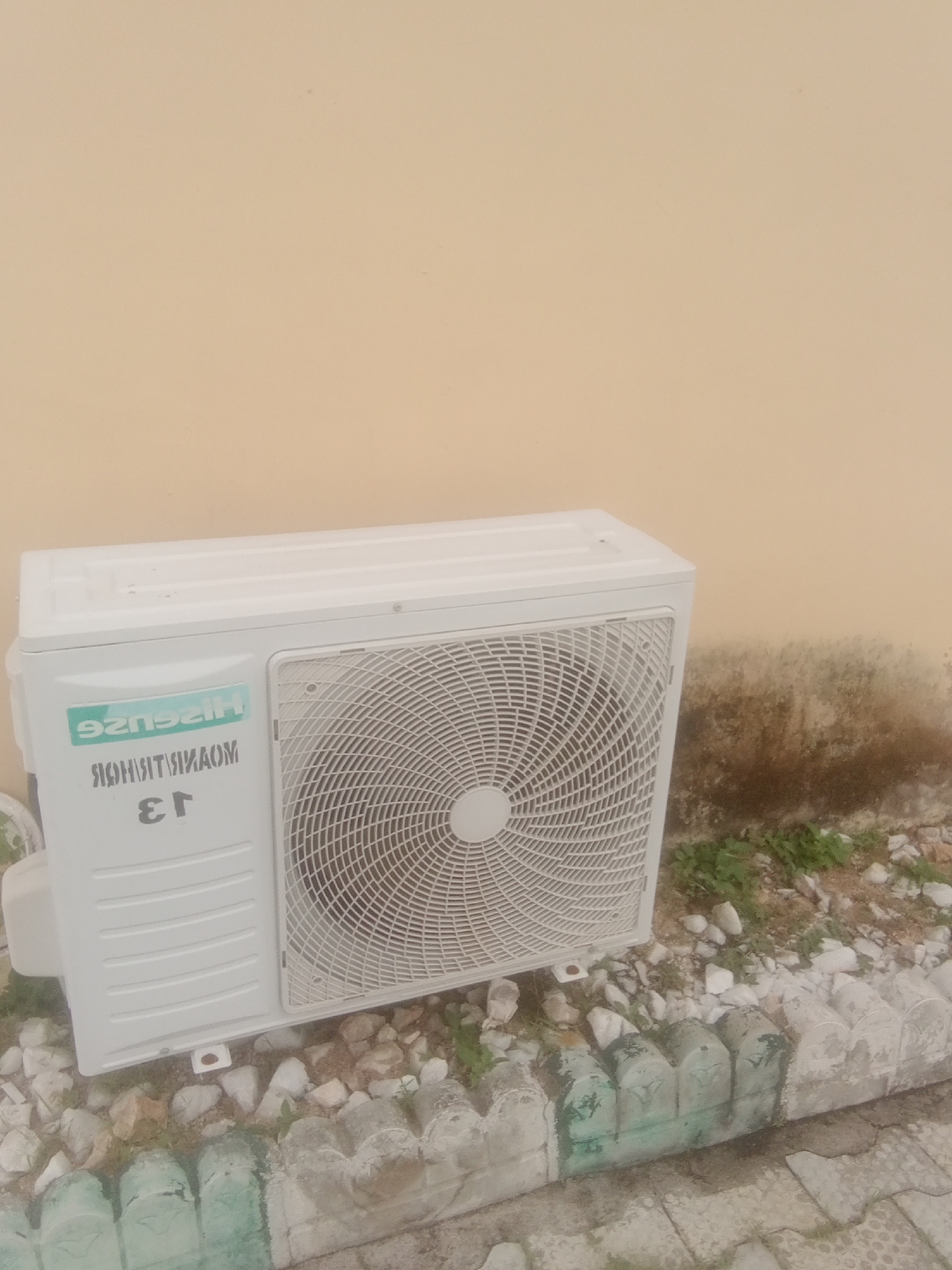 The image shows the outdoor unit of a Hisense air conditioner. The unit is white and has a large fan grill on its side. There is text on the unit that reads "HISENSE" and "ER 13". The air conditioner is placed outdoors, in front of a beige-colored wall. The ground around the unit is covered with small rocks and some sparse greenery. There are also some decorative concrete border elements visible in the foreground. The scene appears to be outdoors during daylight, though the exact time of day and weather conditions are not discernible. The image was taken in Jalingo, Nigeria, which is a location in West Africa. The overall impression is of a functional outdoor appliance in a somewhat unkempt environment.