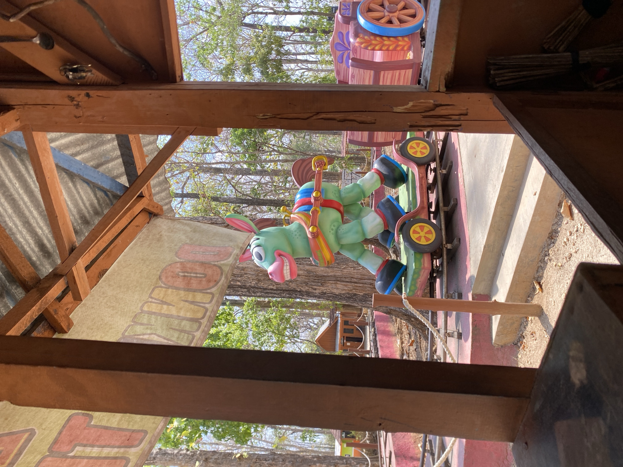 A green, cartoonish donkey-shaped amusement park ride on wheels is positioned on a track outdoors. The donkey has large pink ears, a wide grin, and is adorned with a colorful saddle and harness. It's attached to a wooden train car with decorative purple accents and large wooden wheels. The scene appears to be part of a playground or amusement area, with trees and smaller buildings in the background. The ride is seen through a wooden structure, suggesting it might be under a shelter or part of an indoor/outdoor exhibit. The overall environment is bright and sunny, with dappled light filtering through the trees.