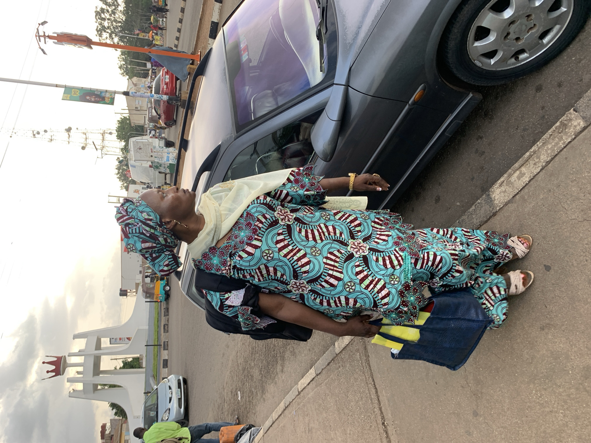 A woman stands next to a dark gray car, looking to her right. She is wearing a blue, green, and red patterned traditional outfit with a matching headscarf and a cream-colored shawl. She has a black backpack on her shoulders and holds a dark blue tote bag with yellow items inside in her left hand. She is also wearing a gold watch and a ring on her right hand. The car has a dark interior and a roof rack. In the background, there is a large white monument with a red crown on top, which is the Barakin Lerawi monument in Nigeria. A silver car, a yellow and blue tuk-tuk, and a white van are visible in the street. The sky is overcast.