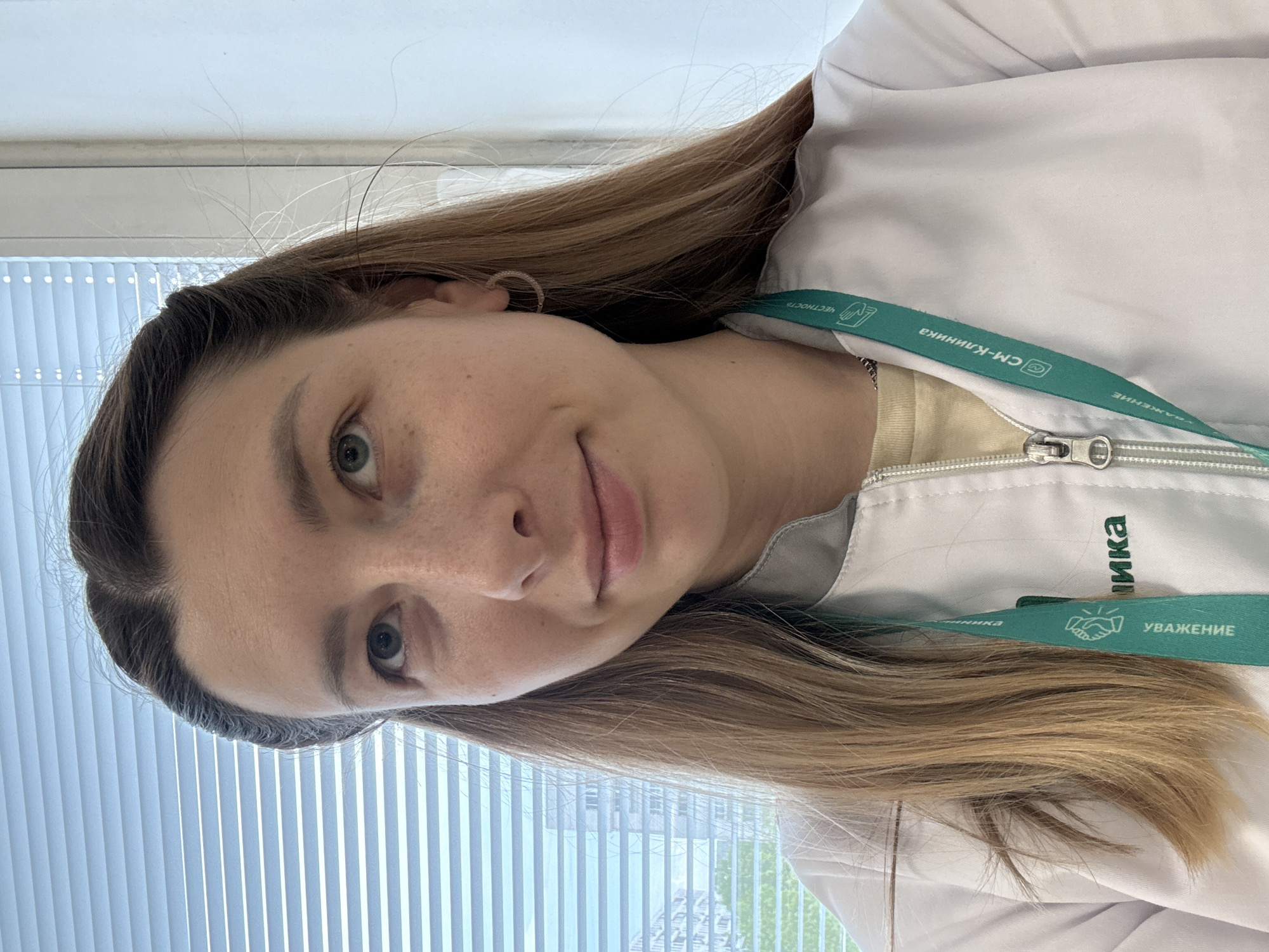 A young woman with light brown hair and fair skin, wearing a white medical coat with a green lanyard, is facing the camera. The lanyard has text in Russian, including "СМ-Клиника" (SM-Clinic) and "Уважение" (Respect). The image appears to be taken indoors, possibly in a medical office or clinic, as suggested by the coat and lanyard. The background shows blinds and a hint of greenery outside the window. Given the clinic's name and the Russian text, the location is likely in Russia, specifically the city of Moscow.