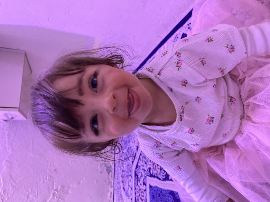 The image displays a young child, appearing to be female and approximately two to four years old, in an indoor setting located in Kandahar, Afghanistan. The child is centrally framed, looking directly at the camera with wide blue eyes and a playful expression, characterized by the tongue slightly extended from the mouth. The child has dark hair, styled with bangs, and prominent long eyelashes. They are dressed in a long-sleeved white top adorned with small pink and blue floral patterns, worn over a light pink, multi-layered tulle skirt. The room is bathed in a distinct purple light. In the background, to the left, a textured, light-colored wall with visible peeling paint or plaster rises from a patterned dark blue rug. To the right, a smoother, light-colored wall is visible. An off-white, wall-mounted fixture, possibly a cabinet or shelf, is situated in the upper right background. The child is positioned on the patterned blue rug.