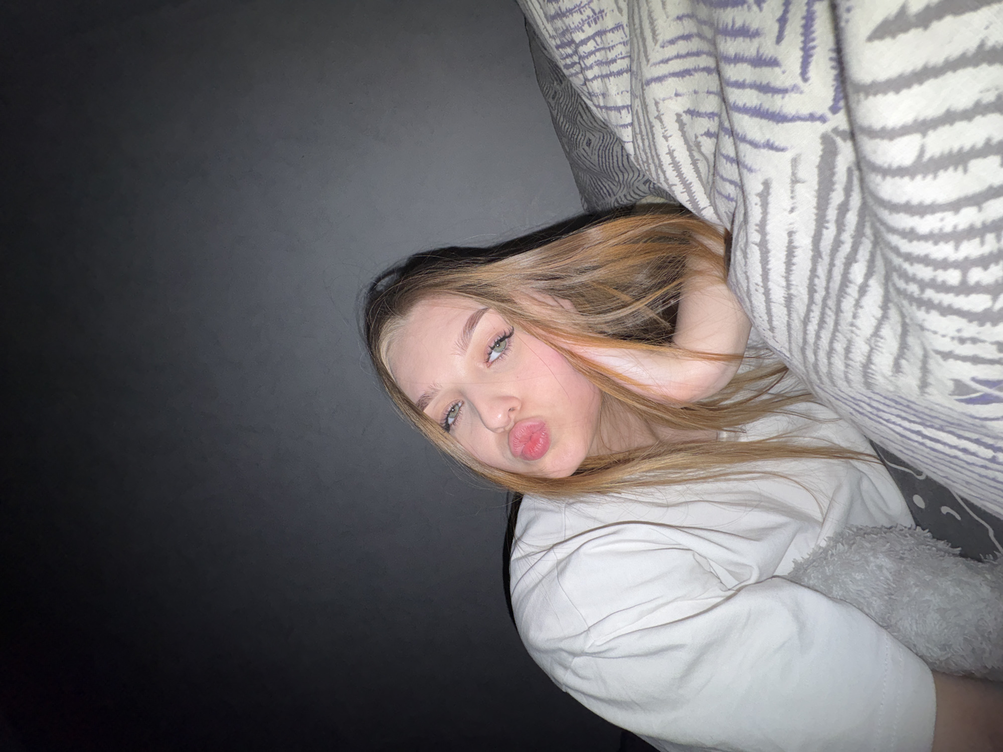 A young woman with light brown hair is lying in bed, partially under a patterned duvet. She is wearing a white shirt and making a kissing face. The background is dark, possibly a wall. The image provides no information about the city of Chaikovsky, Russia.