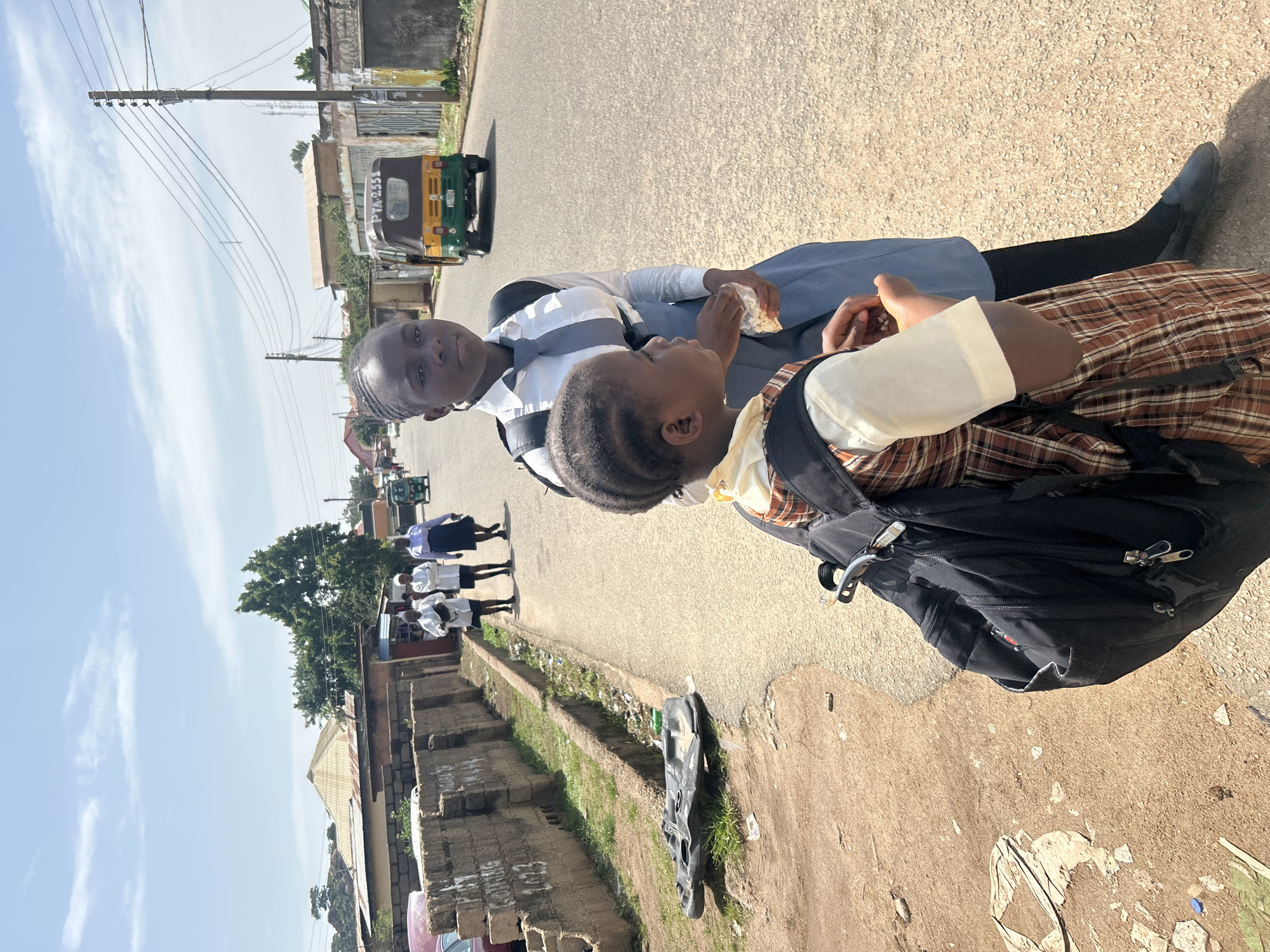 Two young girls, presumably students, are sitting on the side of a paved road in Jos, Nigeria. One girl, on the left, is looking towards the camera with a neutral expression. She is wearing a white school uniform shirt with a grey tie and a blue skirt. The other girl, on the right, is looking down and appears to be eating something white. She is wearing a plaid skirt and a light-colored shirt with a dark backpack. The scene is outdoors, under a clear blue sky with some wispy clouds, suggesting it is daytime. The road is asphalt, and there's a dirt and grass embankment to the left. In the background, a few buildings and utility poles are visible, along with a yellow and green auto-rickshaw (likely a 'Keke Napep'). Other students in similar uniforms can be seen walking in the distance. The overall atmosphere seems casual and everyday. The image has been rotated 90 degrees counter-clockwise.