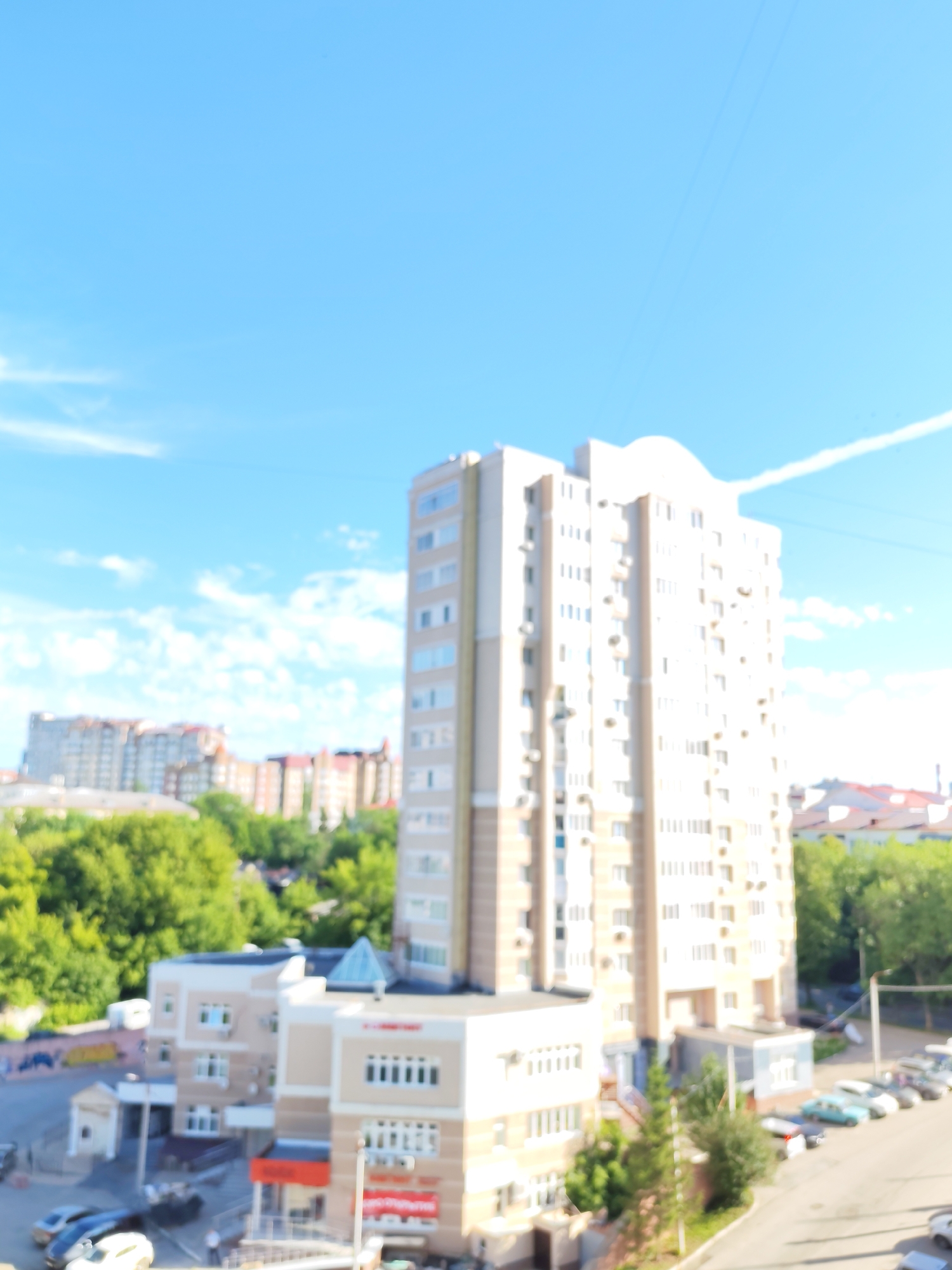 The image captures a daytime scene in a city, likely Ufa, Russia, given the context. A tall, light-colored apartment building dominates the center-right of the frame, featuring numerous windows and balconies. In front of this building, a smaller, multi-story structure with a beige and white facade is visible, possibly a commercial or residential building.
To the left and in the background, other buildings of varying heights are present, some appearing to be apartment complexes with multiple windows and balconies. Lush green trees are scattered throughout the landscape, interspersed between the buildings, suggesting a relatively urban but green environment. The sky is a bright blue with scattered white clouds, and a thin white contrail from an aircraft stretches across it.
A paved road runs along the bottom right of the image, with several parked cars of various colors visible. The image appears to be taken from an elevated perspective, looking down on the scene. The overall impression is of a typical urban residential area under a clear sky.