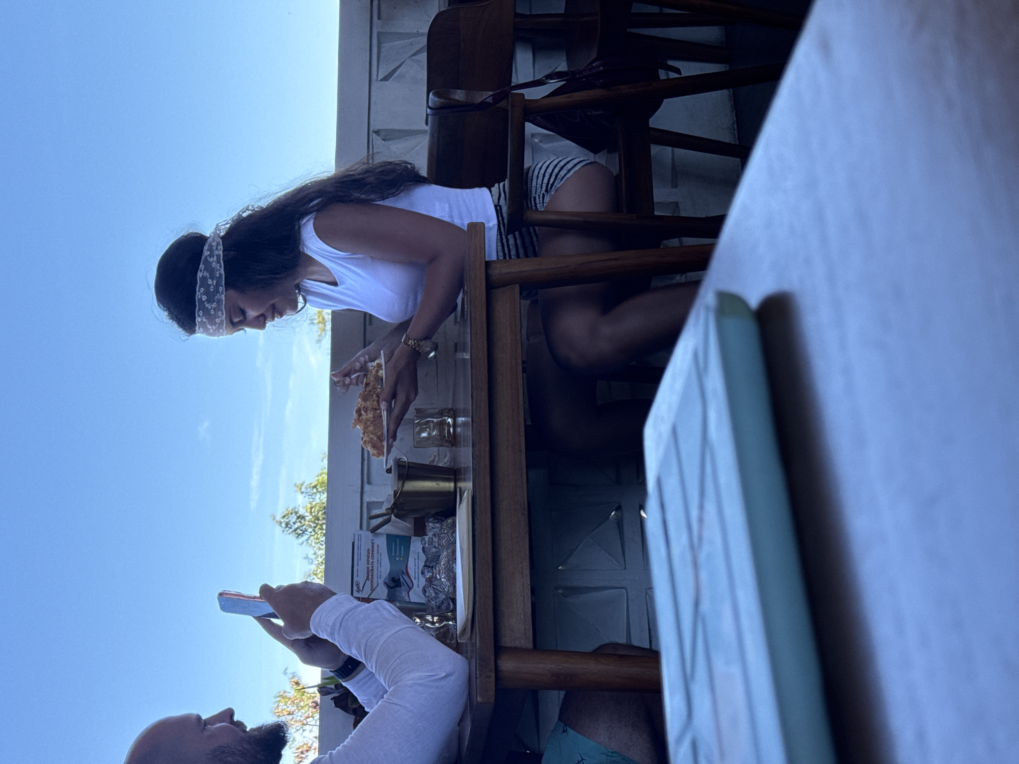 A man with a shaved head and beard is taking a photograph of a woman with long, dark hair wearing a white top and striped shorts with a mobile phone at a restaurant in Kuta Selatan, Indonesia. The woman is seated and eating. There is food, glasses, and a menu on the table. The sky is visible in the background. The building behind them has a textured wall.