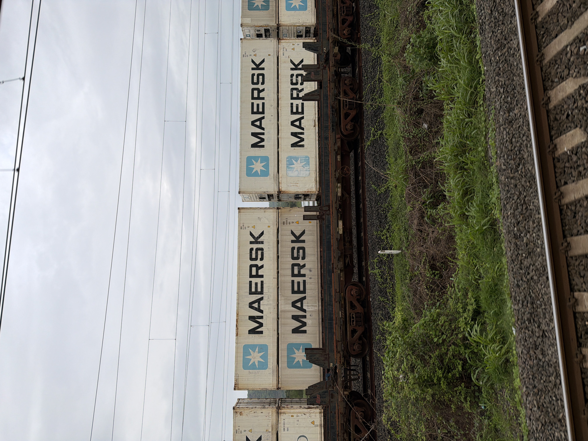 The image shows a train with multiple Maersk shipping containers stacked two high on flatbed railcars. The train is on tracks surrounded by lush green vegetation and some rocky terrain. Overhead power lines are visible against a cloudy sky. The scene is likely near Saphale, India, given the presence of a train line and the surrounding landscape. There are no people visible in the image, and the train is stationary.