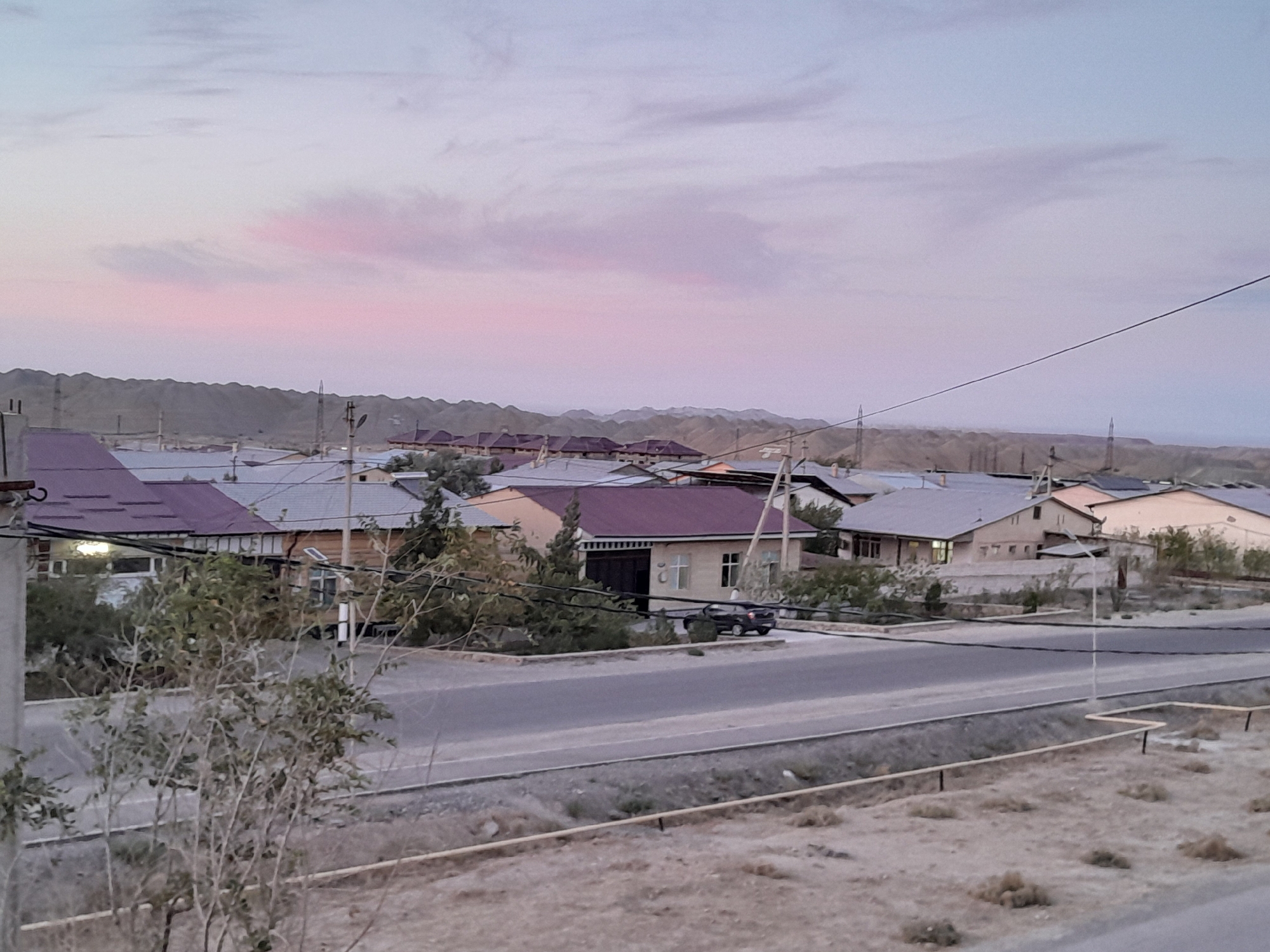 The image captures a view of Uchquduq, Uzbekistan, at dusk. The sky is painted with soft hues of pink and pale blue, with scattered clouds. In the foreground, a dry, dusty embankment with sparse vegetation and a few low-lying plants is visible. A narrow road runs horizontally across the midground, with a paved surface. On the other side of the road, a residential area with several houses is depicted. The houses are single-story structures with varying roof colors, including gray and dark red. Some houses have wooden siding, while others appear to be stuccoed. A dark-colored sedan is parked in front of one of the houses, and a street light stands on the sidewalk. In the background, a range of barren, rolling hills stretches across the horizon, characteristic of the arid landscape surrounding Uchquduq. Power lines are strung across the scene, adding to the infrastructure of the settlement. The overall impression is one of a quiet, rural town at the end of the day.