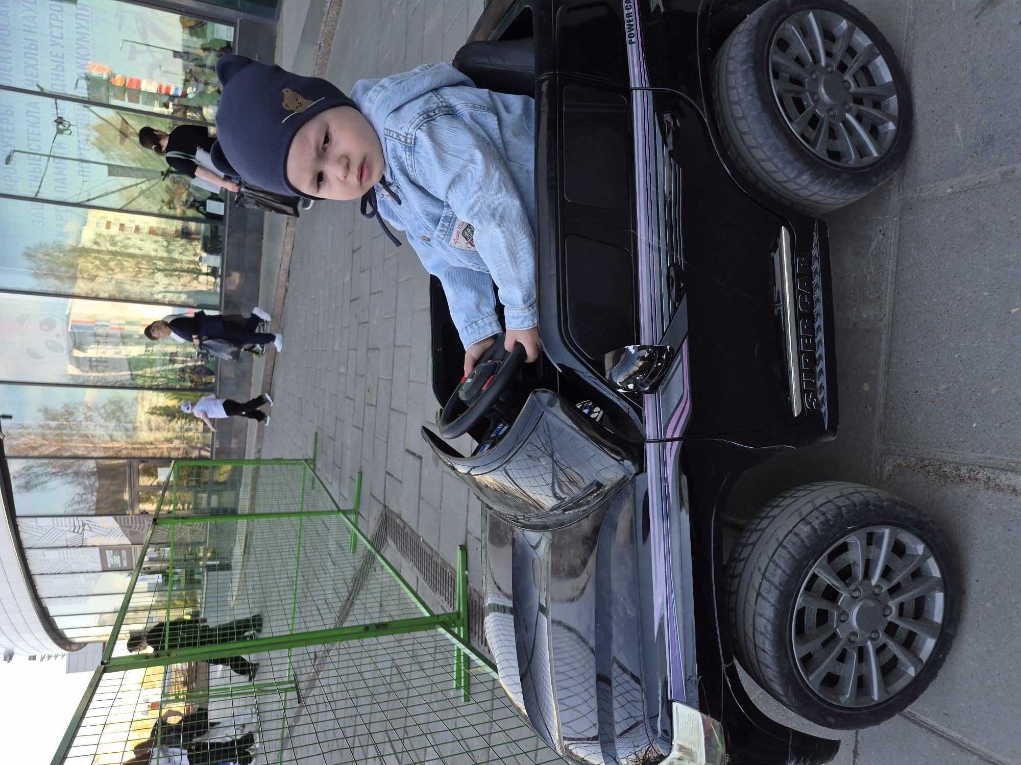 The image shows a young child sitting in a black toy car outdoors. The child is wearing a blue denim jacket and a dark blue hat. The car is positioned on a paved surface, likely a sidewalk, next to a temporary green metal fence. In the background, a building with reflective windows is visible, reflecting people and trees. The text "SUPER CAR" is visible on the side of the toy car. Text in Russian is visible on the building windows "ИРОСКУТЕРЫ ЛЕКТРОСАМ ЗАЩИТНЫЕ СТЕКЛА ЧЕХЛЫ НАУС КАРТИ ПАМЯТИДНЫЕ УСТРО ПОРТАТИВИ ДЕАККУМУЛЯ". The location is suggested to be Tobolsk, Russia.