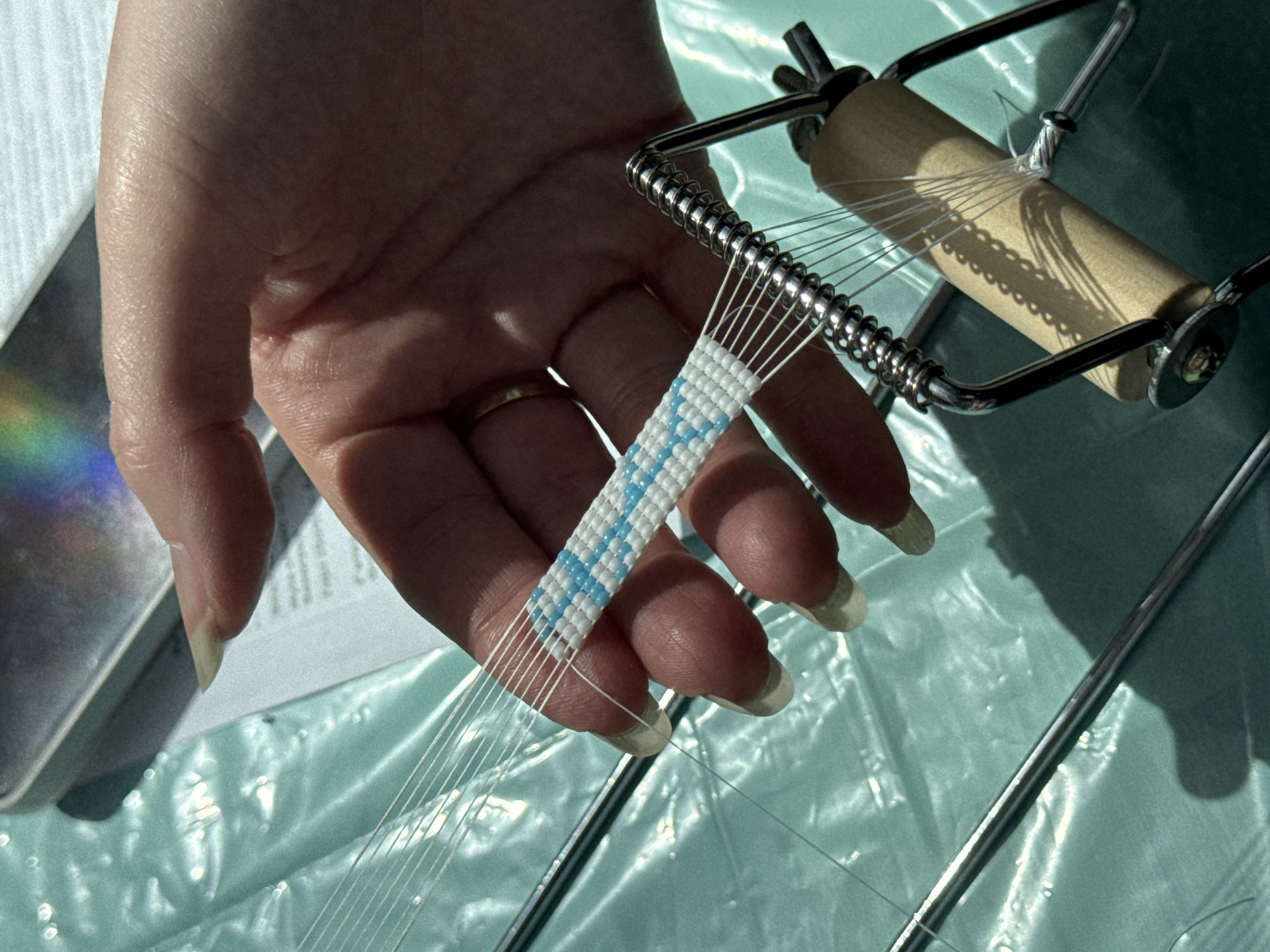 A close-up shot shows a person's hand with long, light-colored fingernails weaving a beaded bracelet on a loom. The bracelet is made of white and light blue beads in a geometric pattern. The loom is a metal device with a wooden spool. The background is a teal-colored surface with a textured, plastic-like finish. A small piece of paper with text is visible under the hand. The lighting suggests a bright, natural light source casting shadows. There is no indication of the location being in Kilkenny, Ireland.