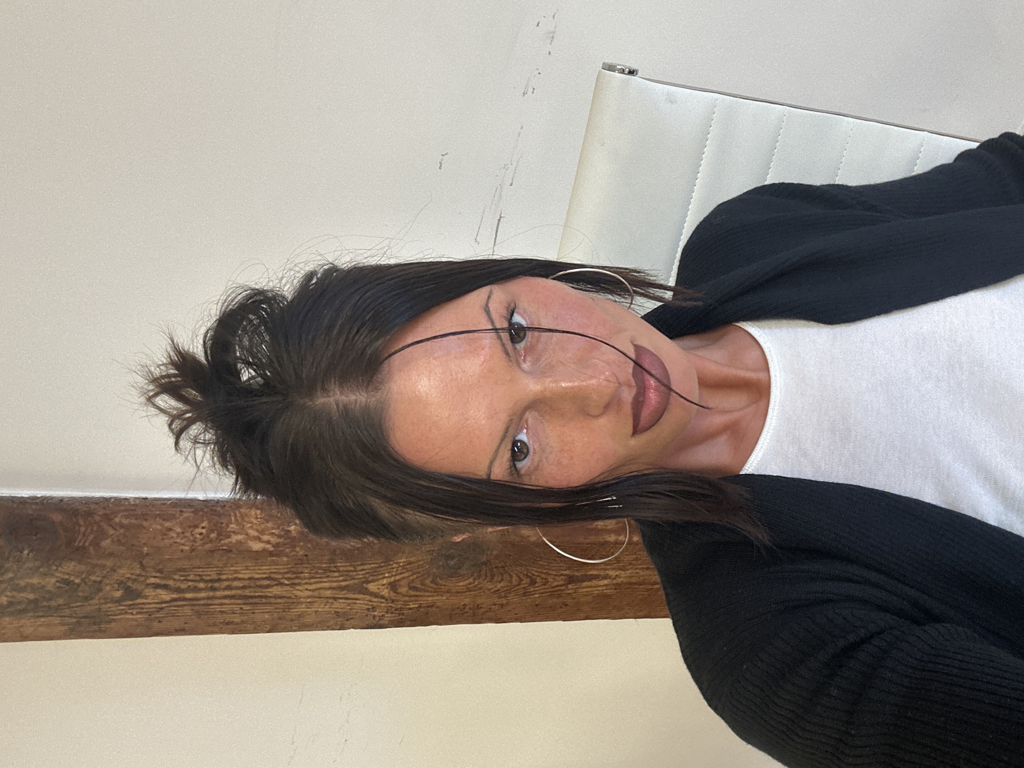 The image displays a close-up portrait of a person, likely female, from the chest up, with the original image rotated 90 degrees clockwise. The person has dark brown hair, styled with some strands falling across the face, and the rest tied up in a high, somewhat tousled bun or ponytail. Their skin is light with visible freckles, and they have dark brown eyes, looking directly at the camera with a neutral to slightly serious expression.
Notable facial details include dramatic, graphic black eyeliner extending from the outer corner of the eyes towards the temples, and dark reddish-brown lipstick with a darker lip liner. Large, silver-toned hoop earrings are visible on both ears.
The person is wearing a plain white top beneath a black, ribbed long-sleeved cardigan.
The background consists of a light beige or off-white wall. A prominent, distressed wooden beam runs horizontally across the lower part of the frame, behind the person's shoulder and chest. To the right of the person's head, part of a white upholstered chair with visible stitching is visible. The scene suggests an indoor setting, possibly a room with a mix of modern and rustic elements. There is no discernible activity other than posing for the photograph. No text is visible in the image.